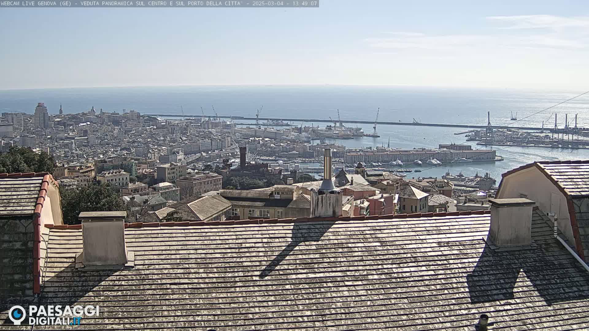 A panoramic view of Genoa, Italy, on a clear, sunny day shows the city's harbor, buildings, and a section of the Mediterranean Sea.