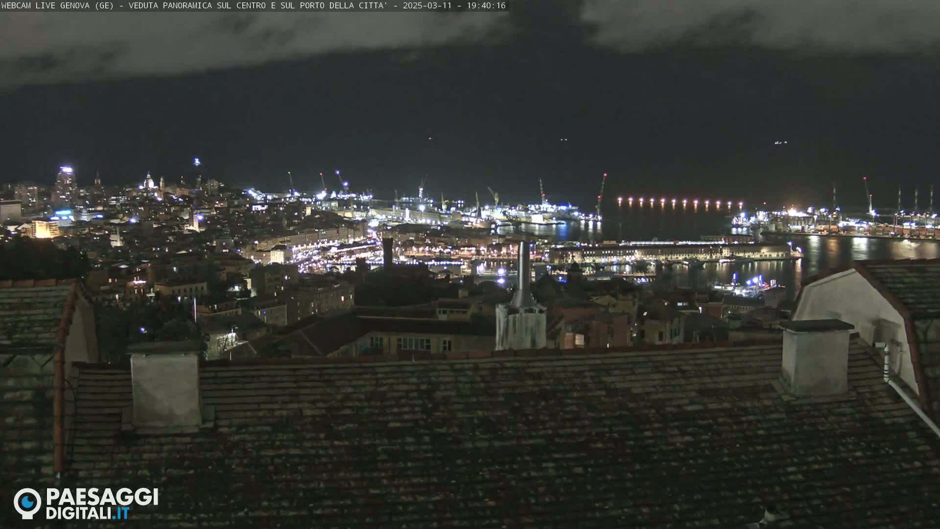 A nighttime panoramic view of a city harbor, illuminated with lights, under a mostly cloudy sky.