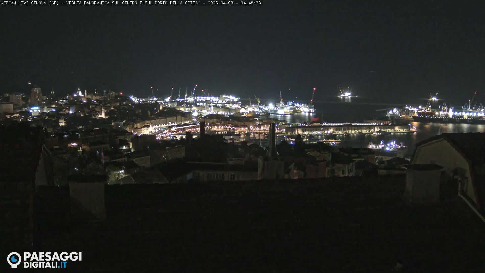 A nighttime, panoramic view of a city's illuminated harbor and buildings, seen from a higher vantage point.