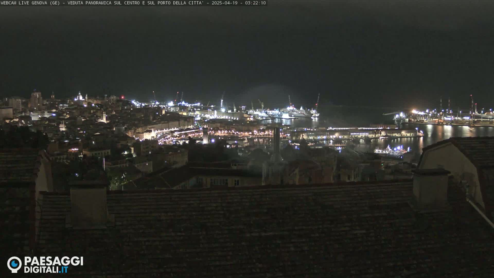 A nighttime, panoramic view of a city's harbor and illuminated buildings, seen from a higher vantage point.