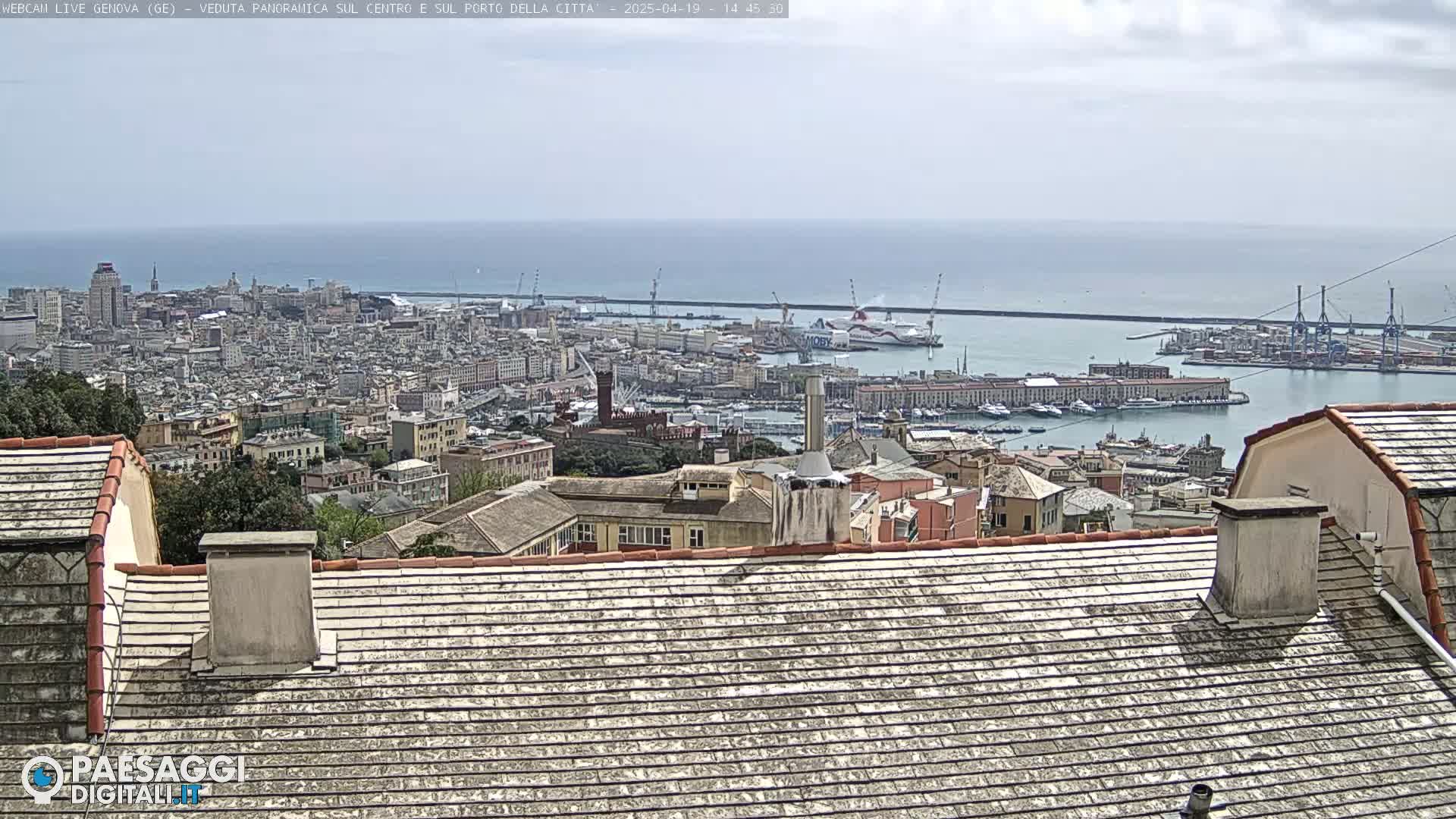 A panoramic view of a city and harbor on a partly cloudy day shows a vast cityscape extending to a body of water with numerous ships and a breakwater.