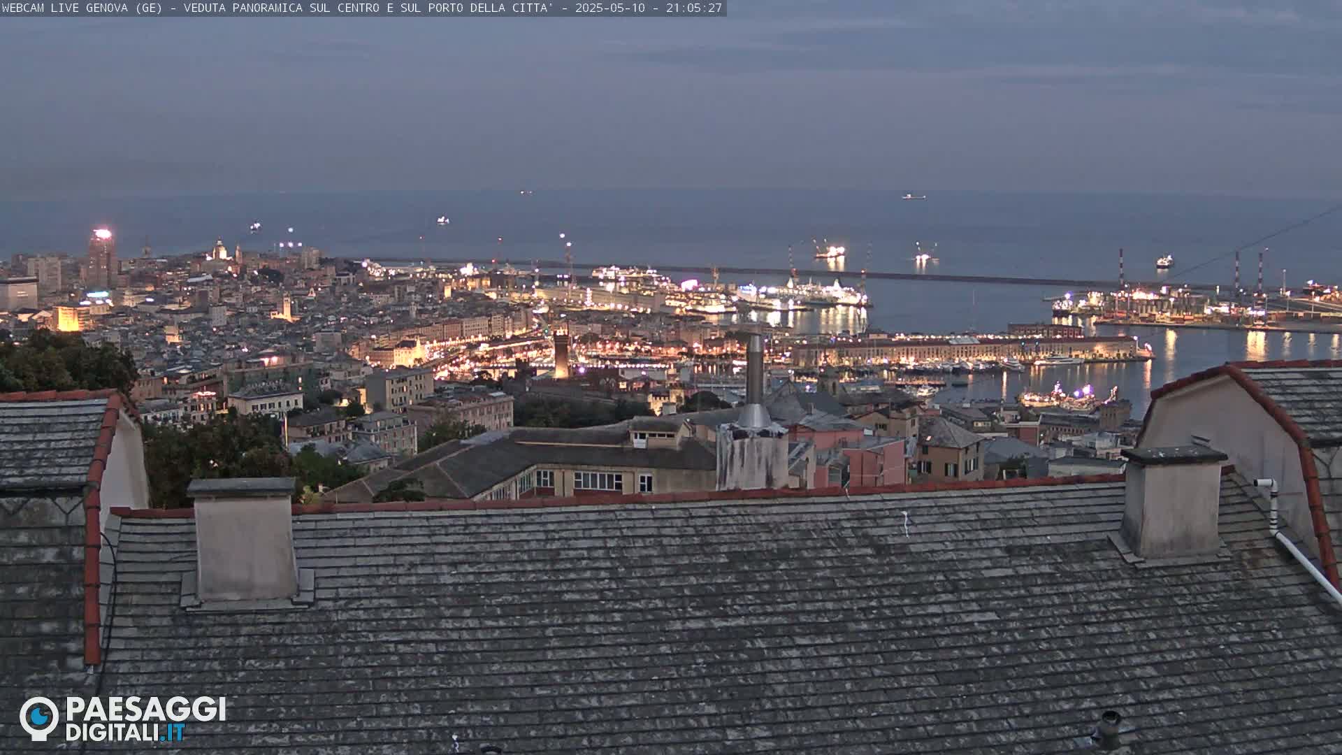 A nighttime, panoramic view of a city's harbor and cityscape, illuminated by lights, extends to a dark, calm sea.