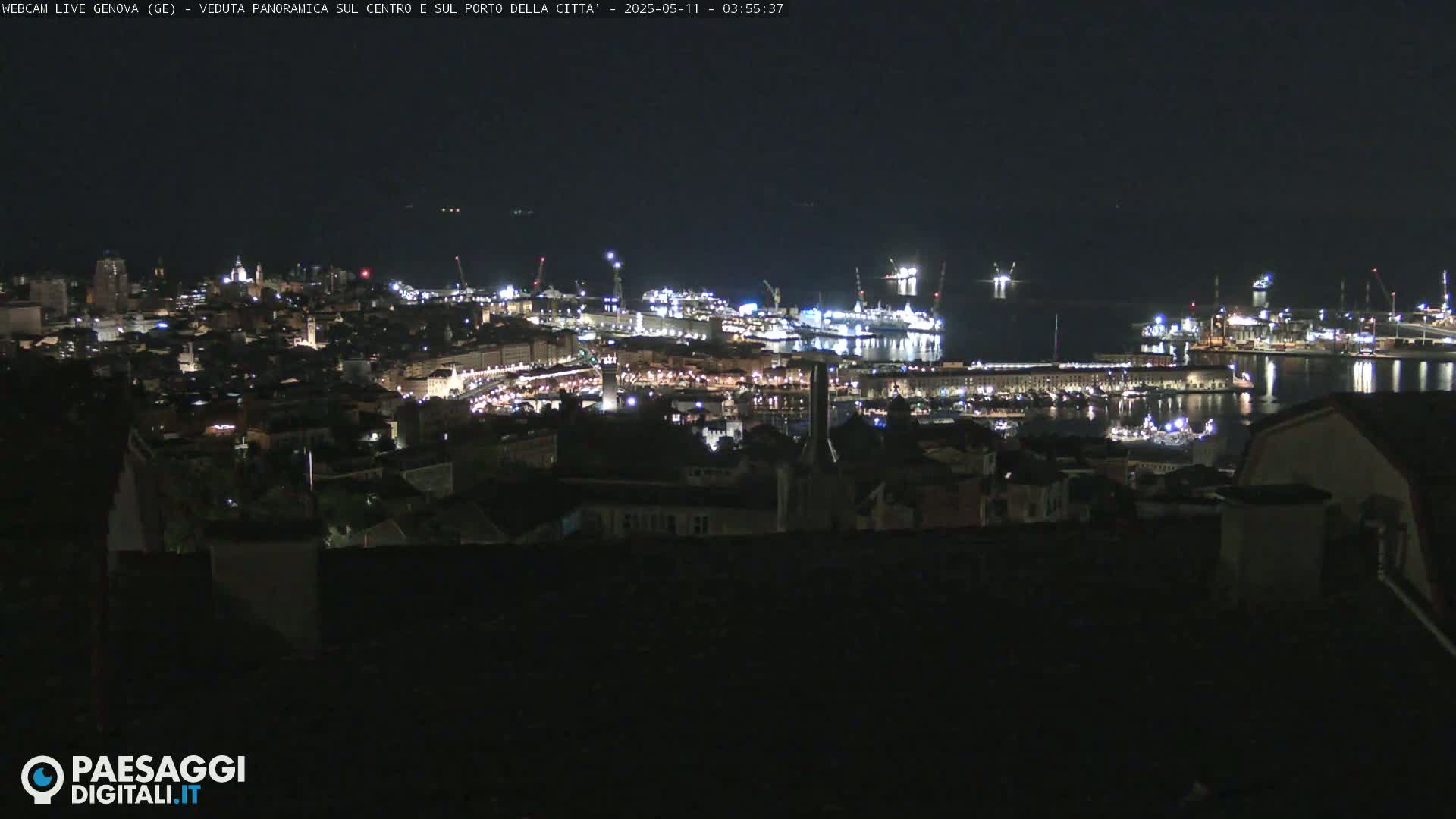 A nighttime, panoramic view of a city's harbor and illuminated cityscape, with a dark body of water reflecting the city lights.