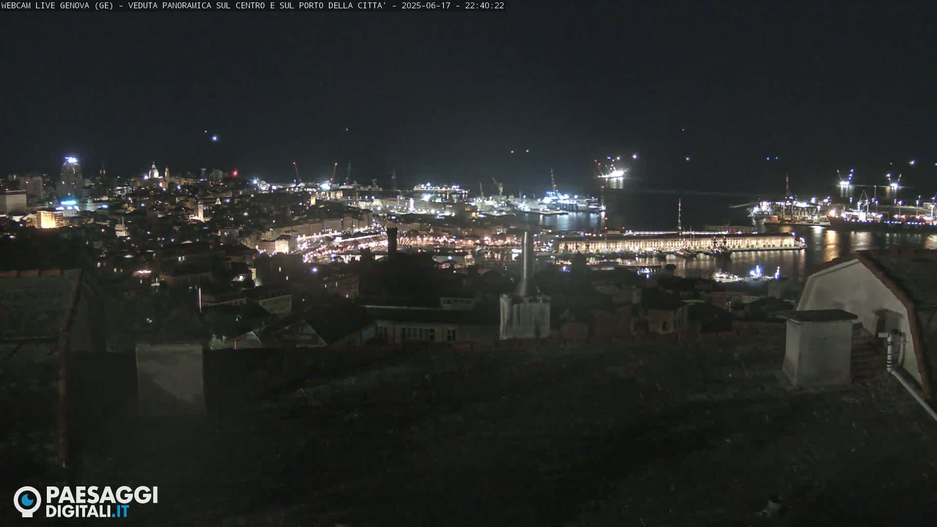 A nighttime, panoramic view of a city's harbor and illuminated cityscape, with several ships and cranes visible in the port.