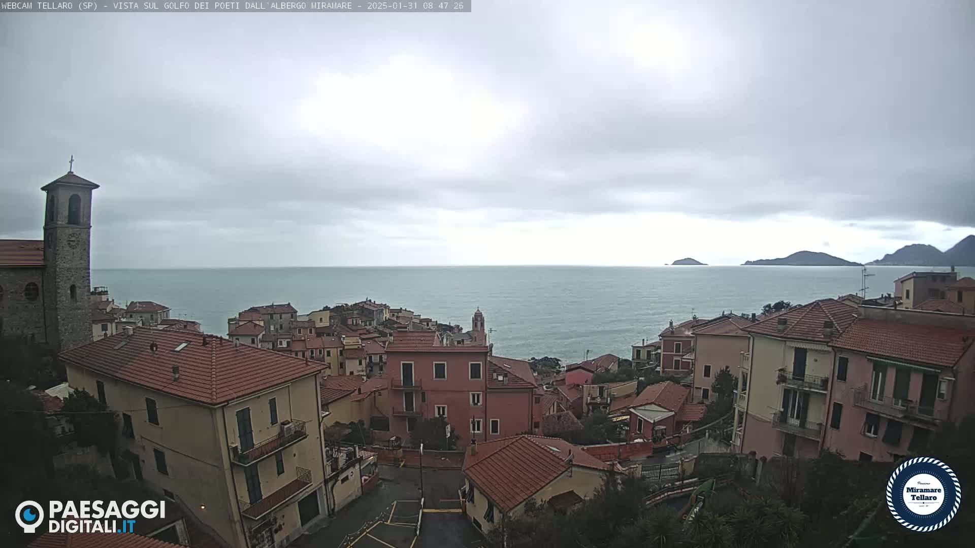 A coastal town with red-tiled roofs overlooks a calm sea under a cloudy sky.