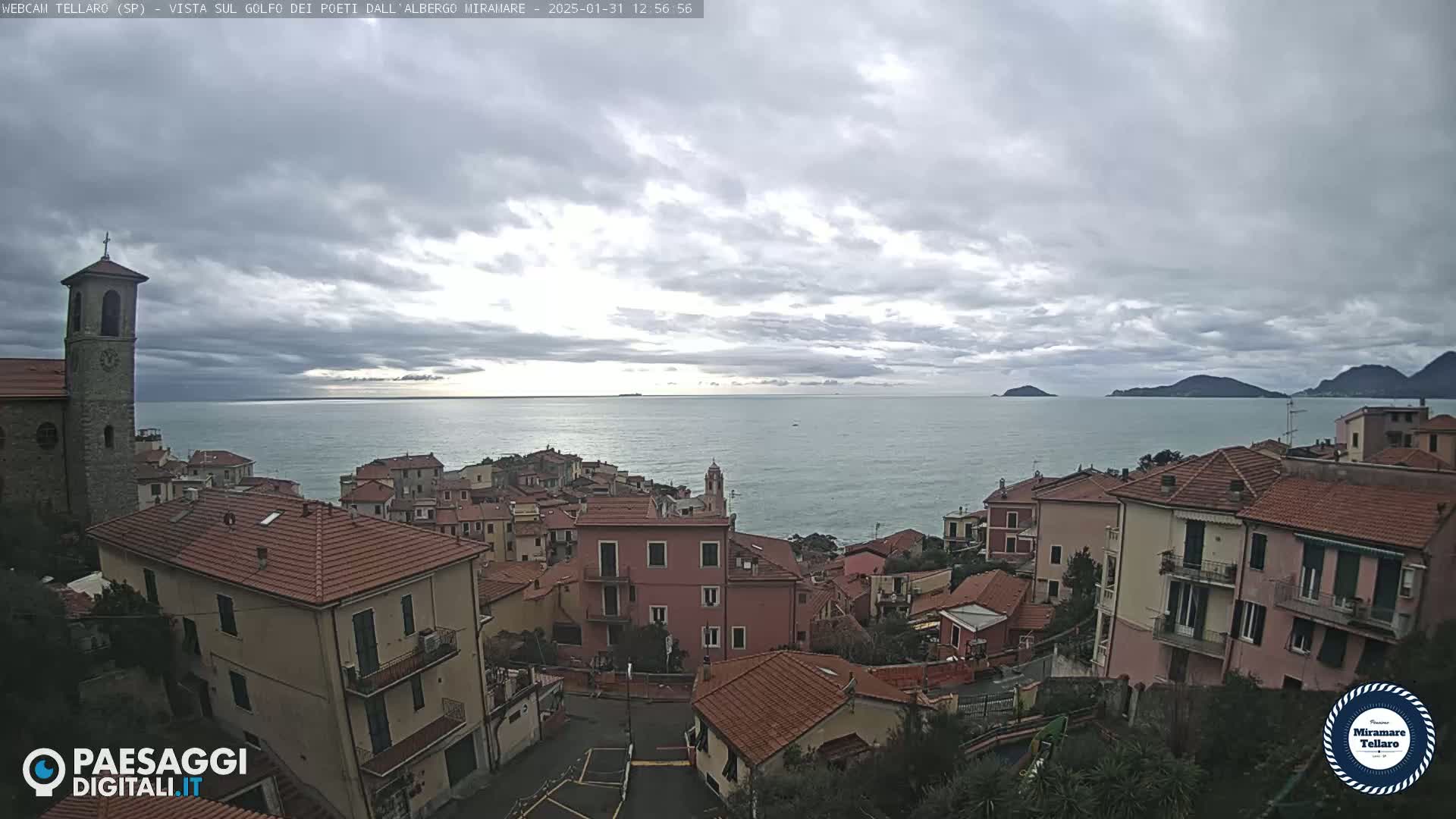 A coastal town with red-tiled roofs overlooks a calm sea under a cloudy sky.