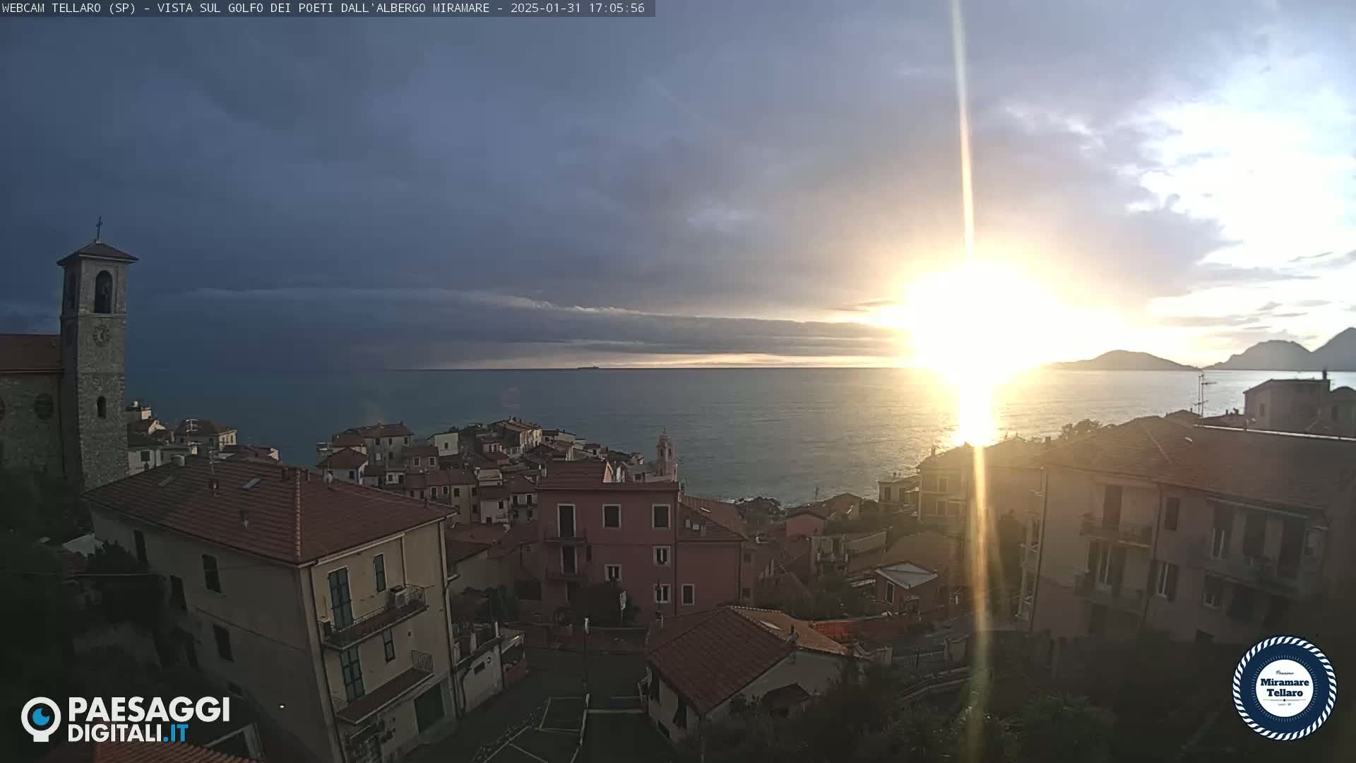 A coastal town with red-tiled roofs sits on a hillside overlooking a calm sea at sunset under a partly cloudy sky.
