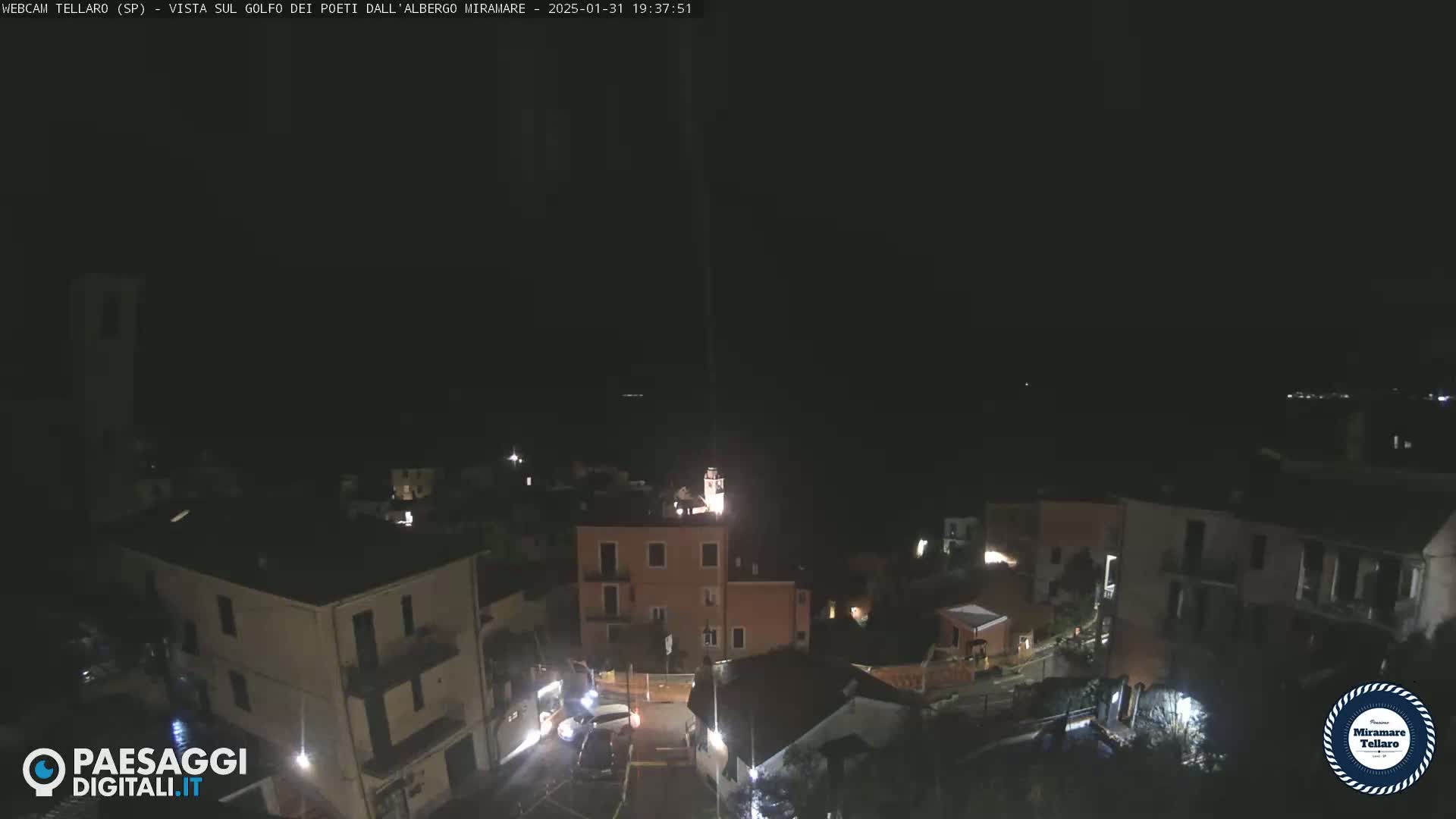 A nighttime view of a small town nestled on a hill overlooking a dark body of water under a clear, dark sky.