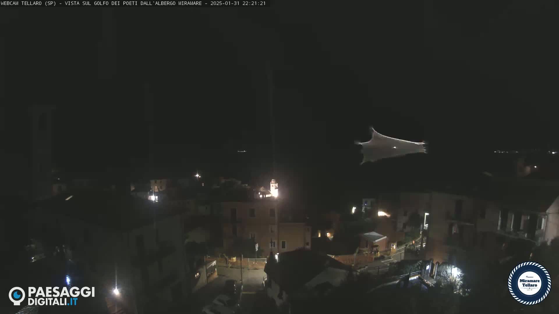 A nighttime aerial view of a small town, possibly coastal, with a large, bright, sheet-like object floating in the dark sky above.