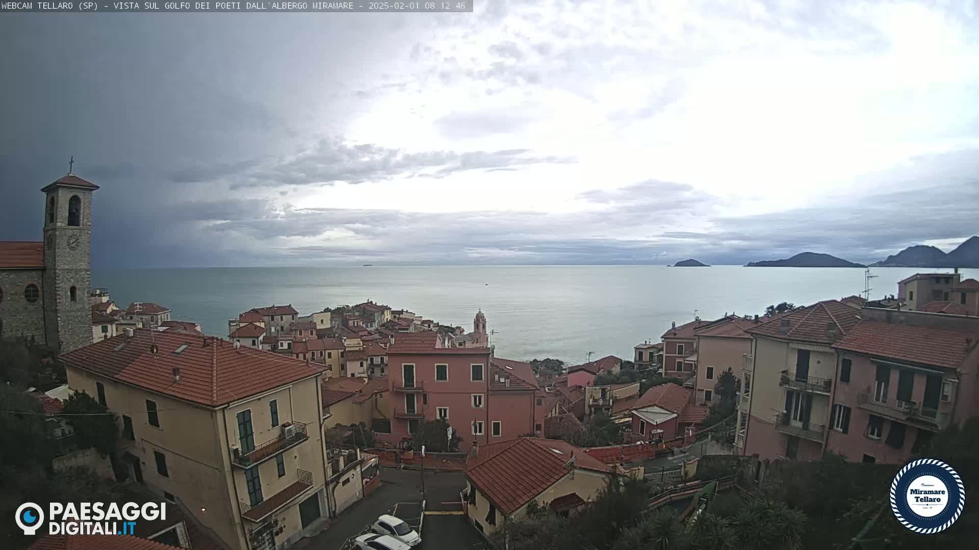 Overcast skies above a coastal town with red-tiled roofs, overlooking a calm sea and distant islands.