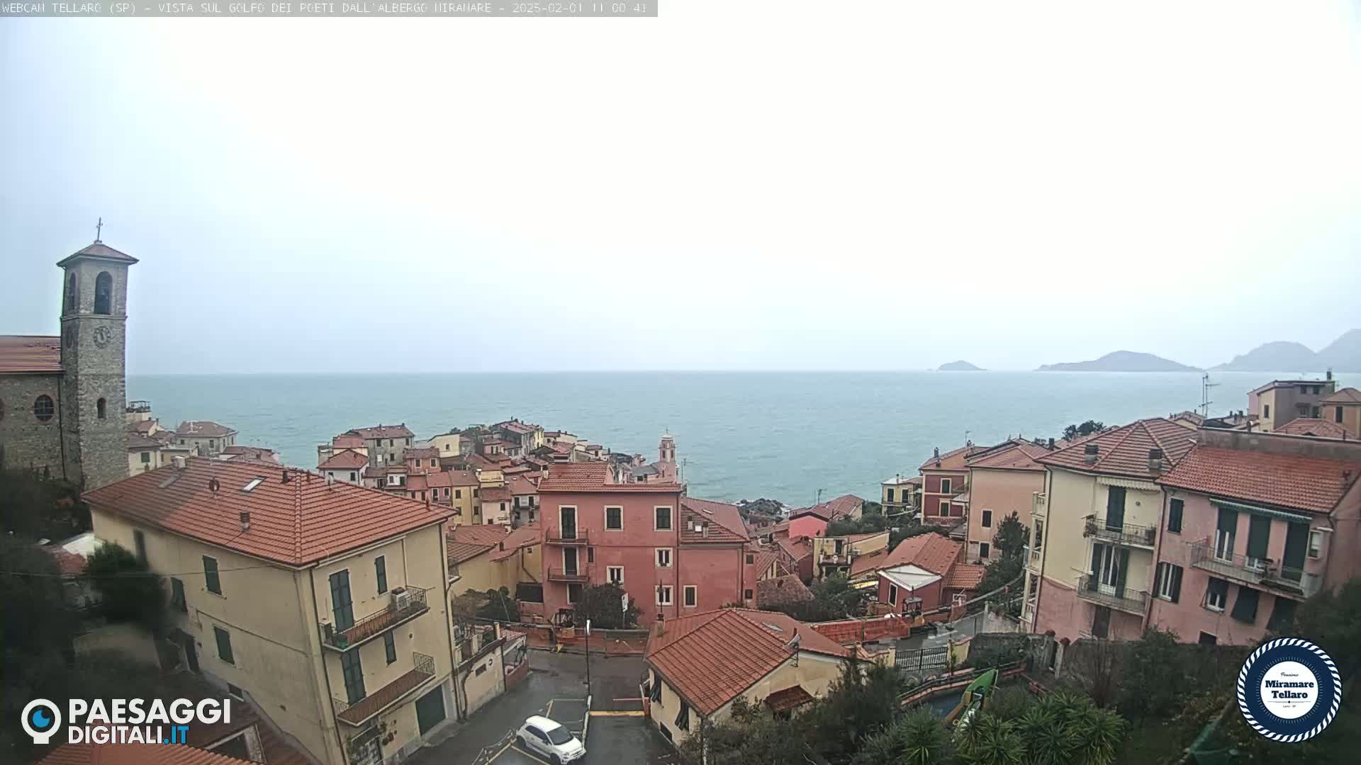 A coastal town with red-tiled roofs and a church tower overlooks a calm sea under an overcast sky.