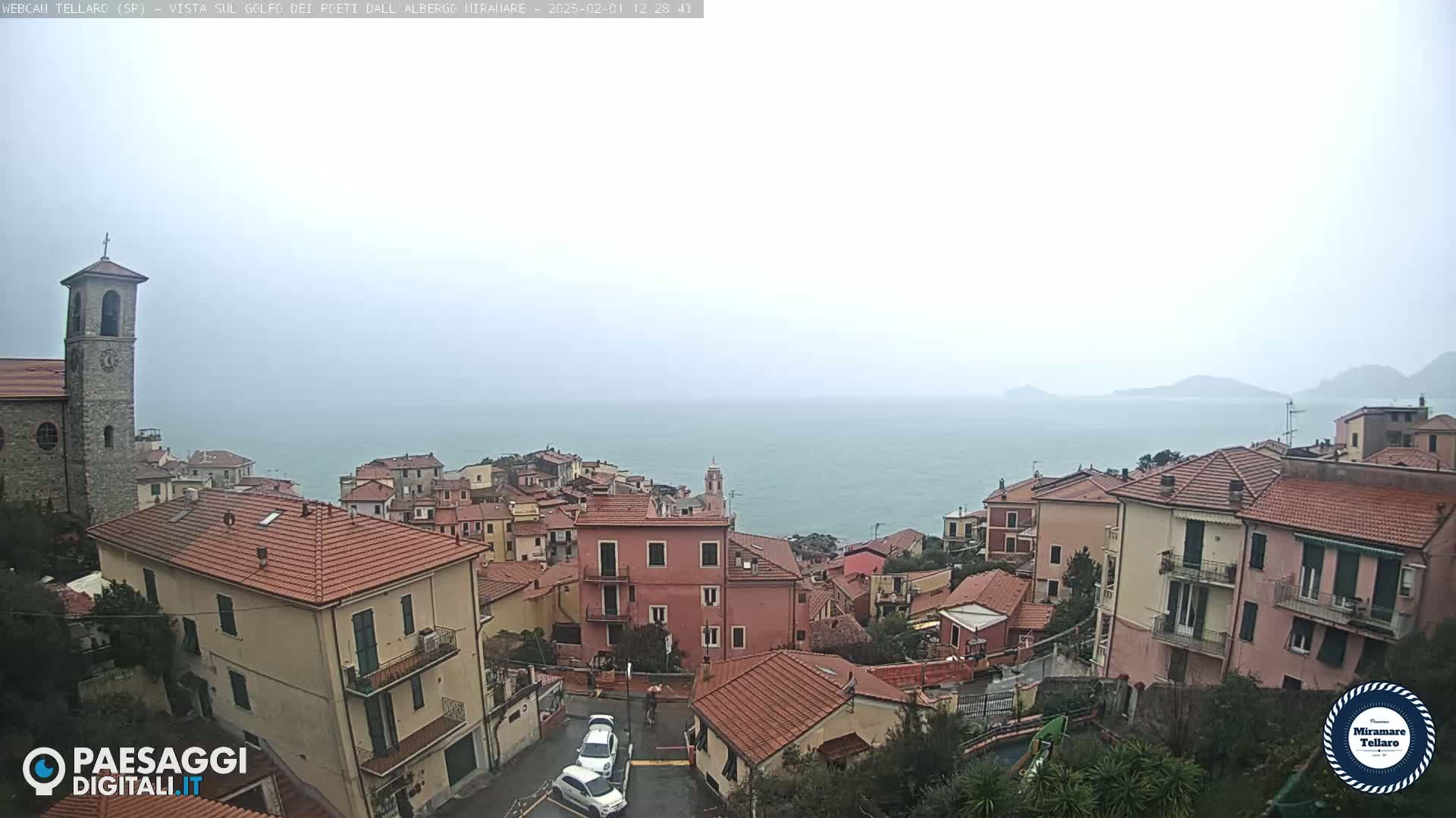 A coastal town with red-tiled roofs overlooks a calm sea under an overcast sky.