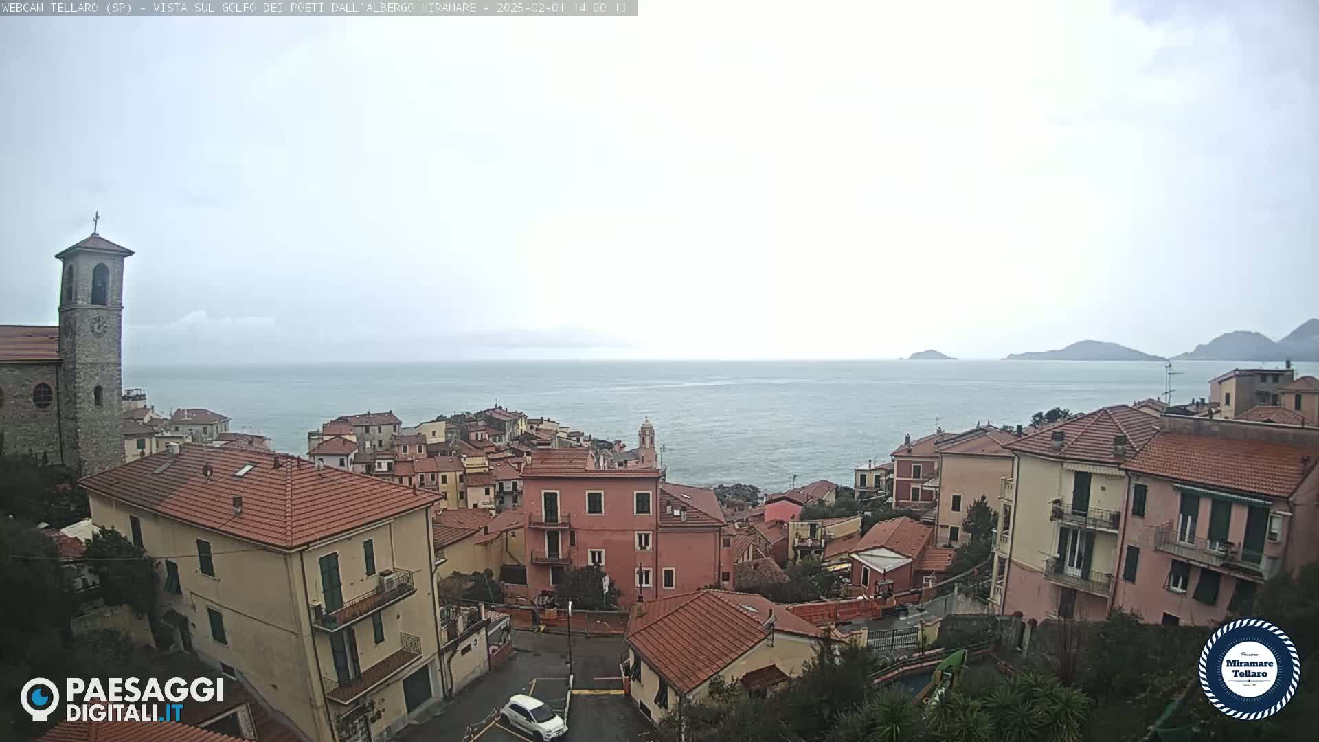 A coastal town with red-tiled roofs and a church tower overlooks a calm sea under an overcast sky.