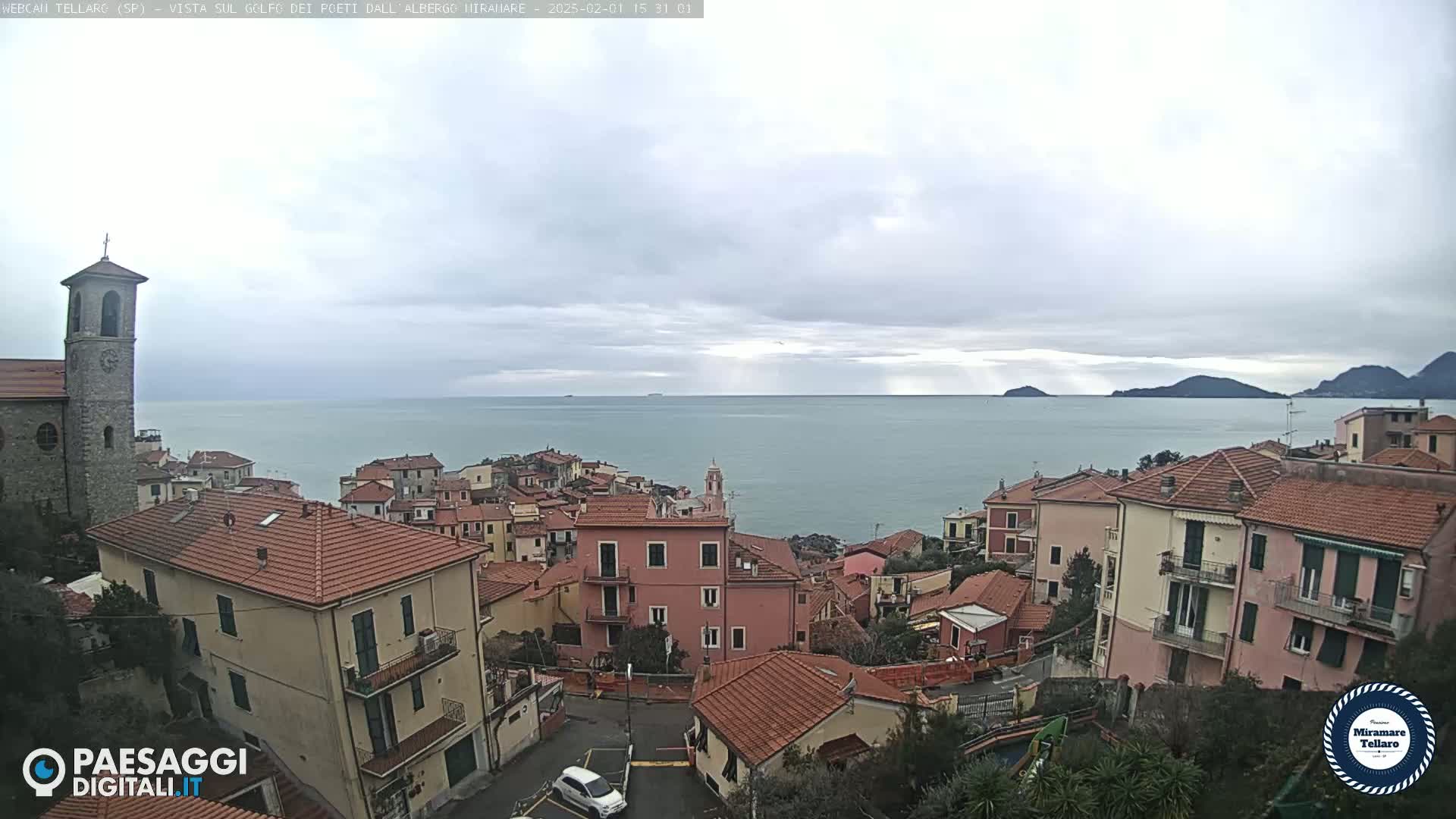 A coastal town with red-tiled roofs and a church steeple overlooks a calm sea under an overcast sky.