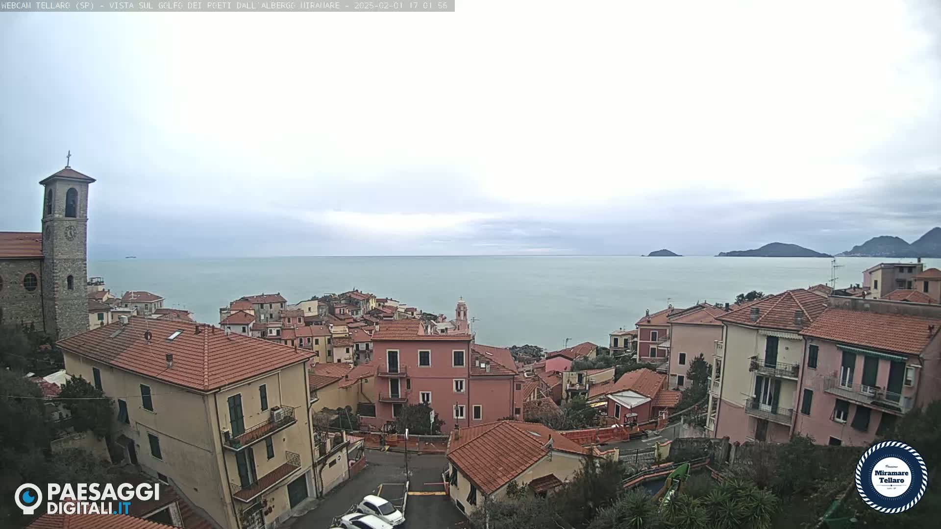 A coastal town with red-tiled roofs and a church overlooks a calm sea under a cloudy sky.
