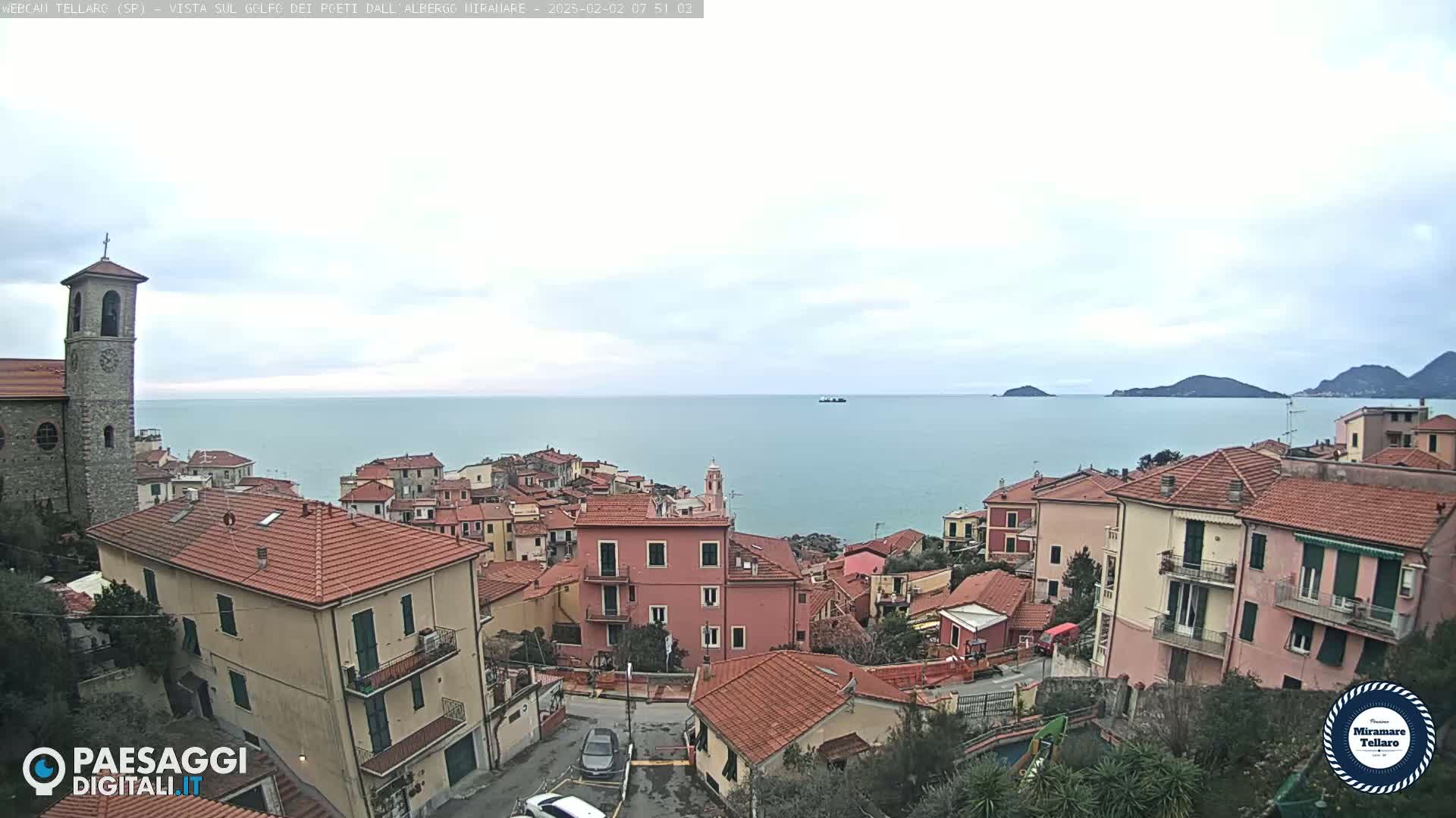 A coastal town with red-tiled roofs and a church steeple overlooks a calm sea under a cloudy sky.