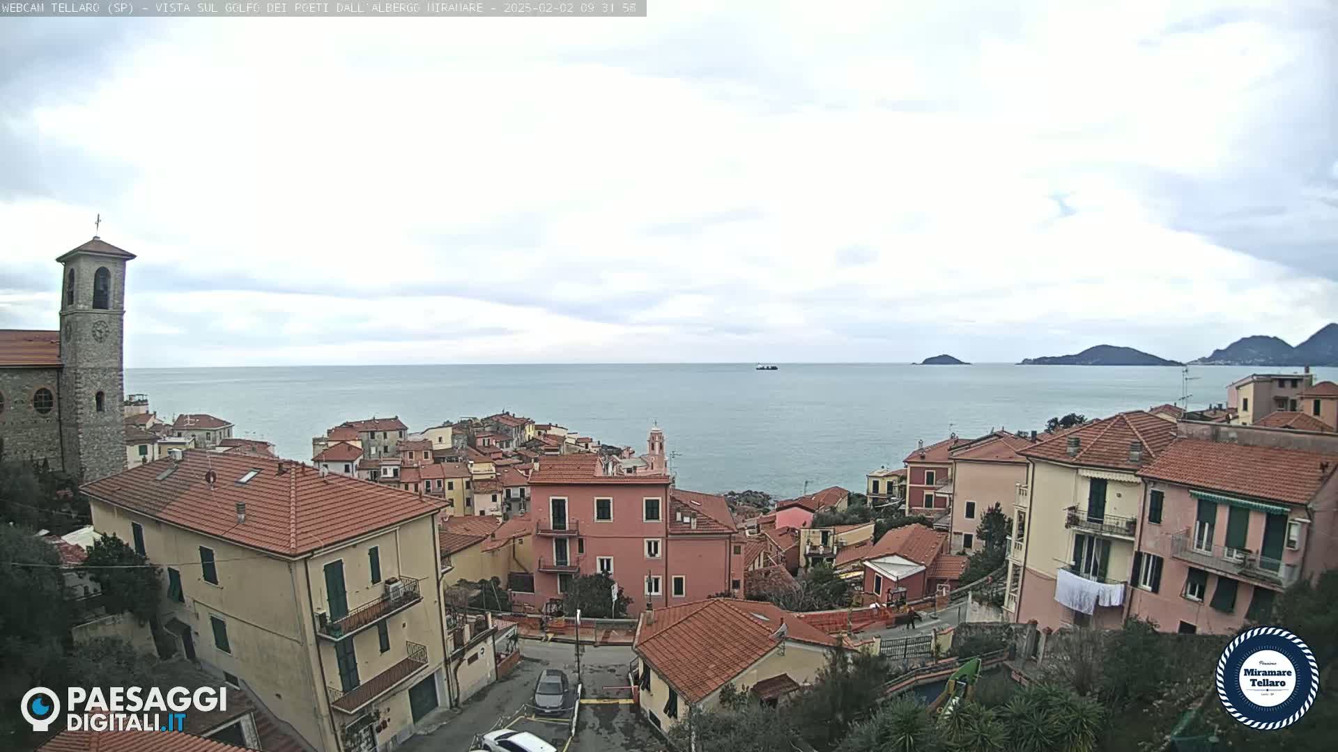 A coastal town with red-tiled roofs and a church overlooks a calm sea under a mostly cloudy sky.