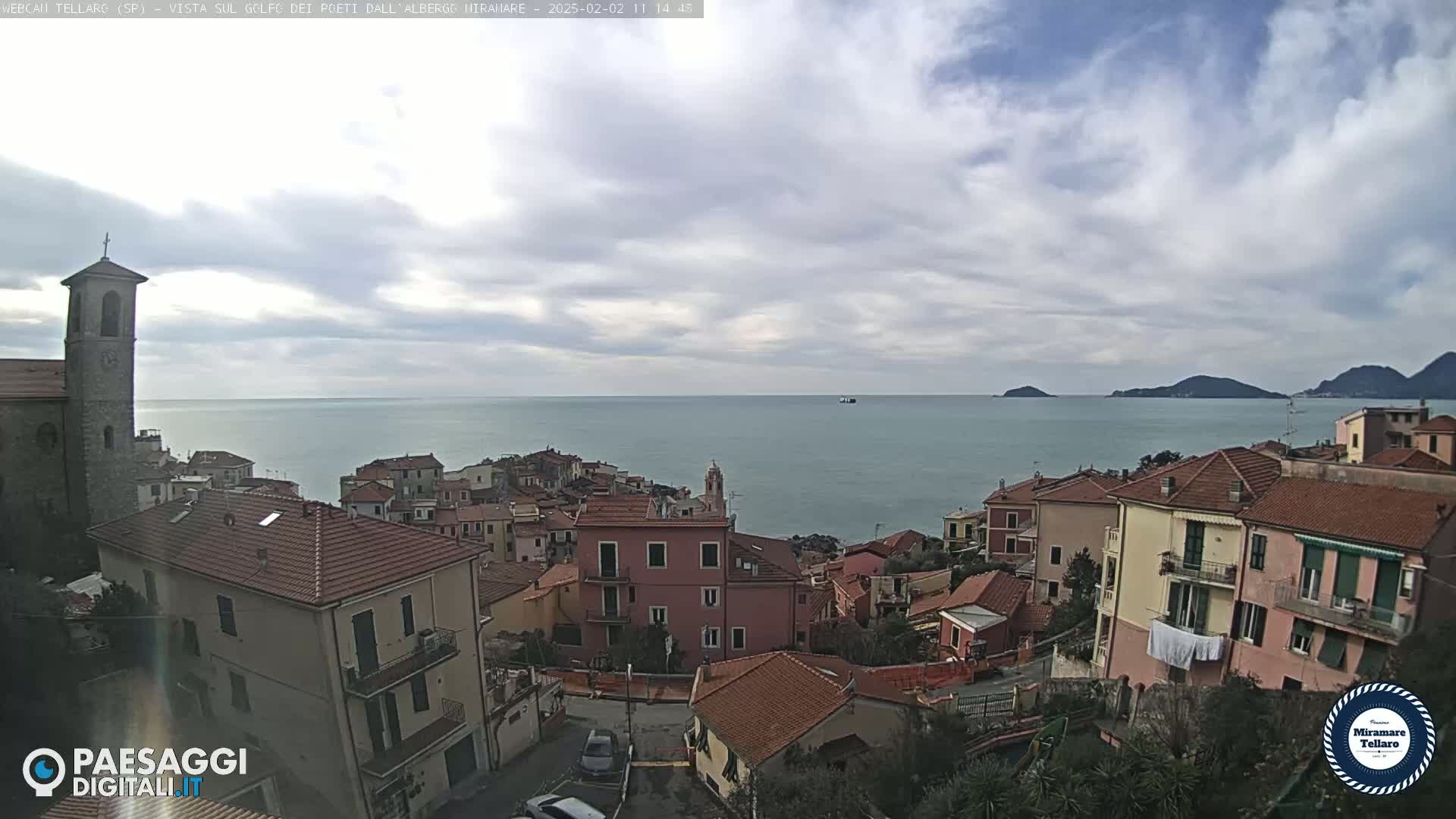 A coastal town with red-tiled roofs and a church steeple overlooks a calm sea under a partly cloudy sky.