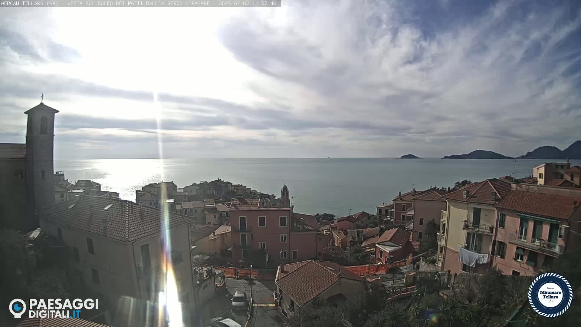 A coastal town with red-tiled roofs and a church steeple overlooks a calm sea under a partly cloudy sky.