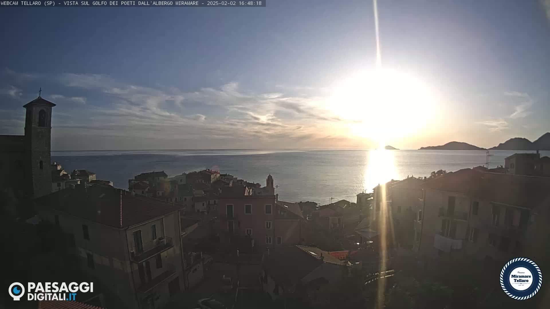 A coastal town with a church steeple is shown at sunset over a calm sea with hazy sunlight.