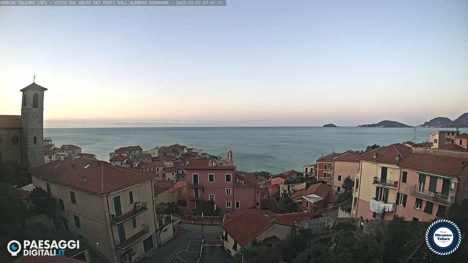 A coastal town with red-roofed buildings overlooks a calm sea under a clear, pale sky at dawn.