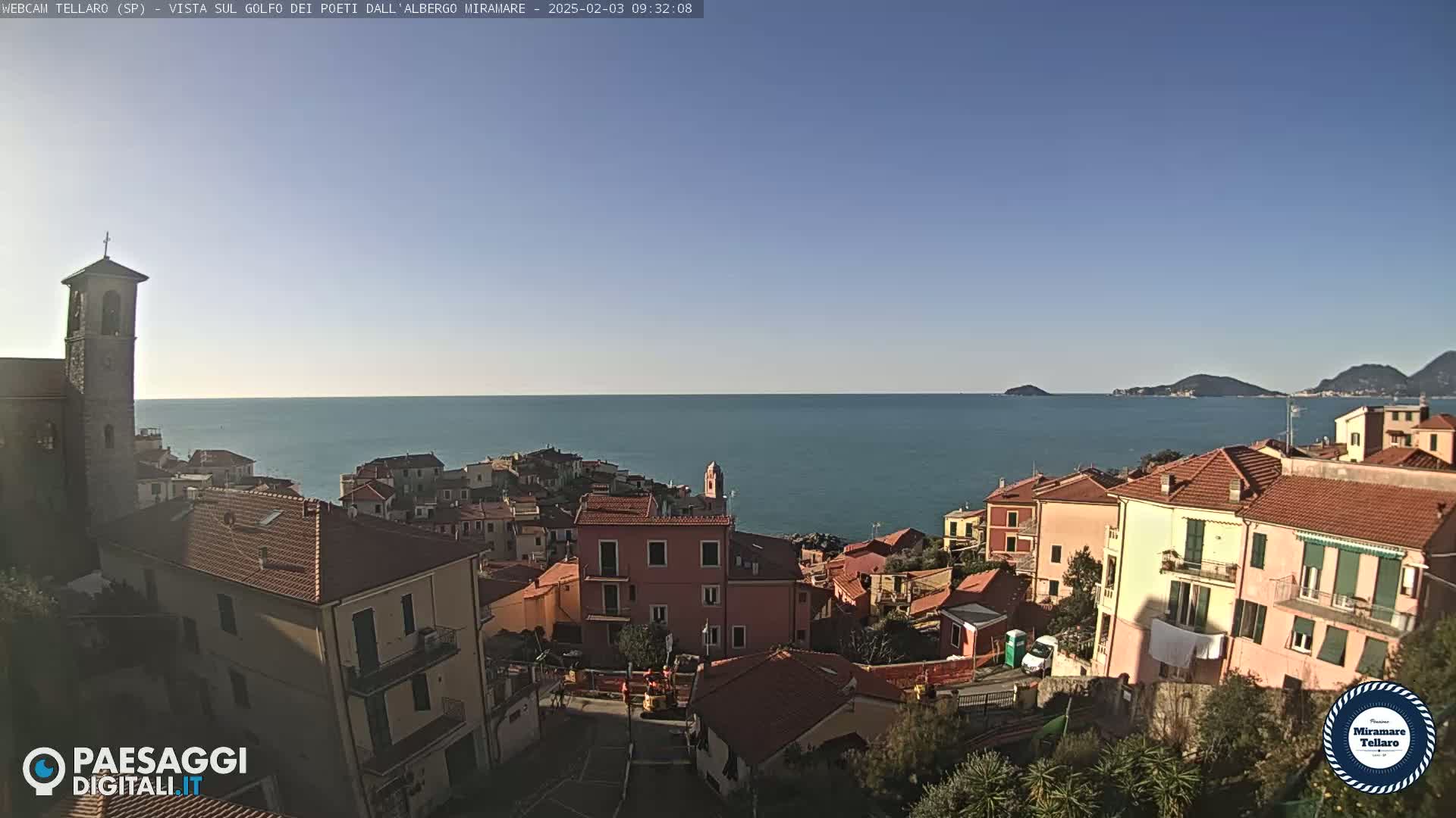 A coastal town with red-tiled roofs and a church steeple overlooks a calm blue sea and distant hills under a clear blue sky.