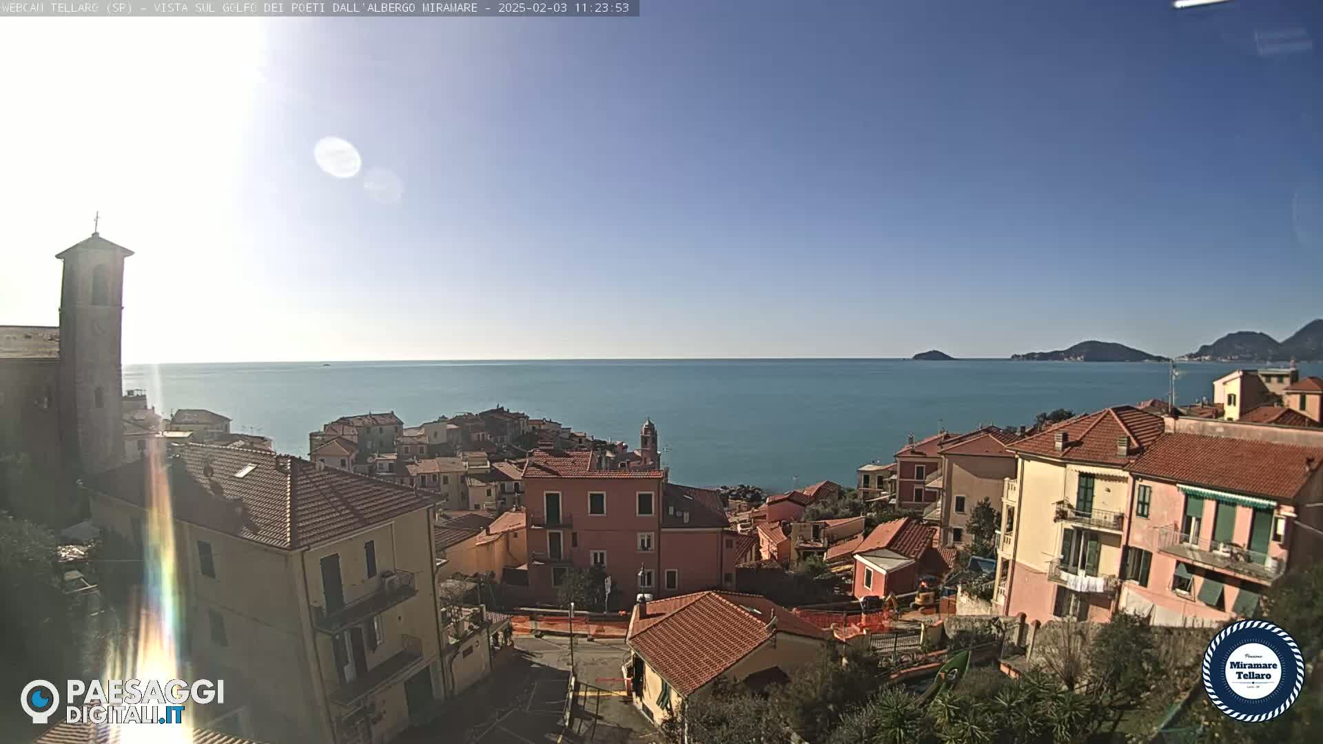 A sunny day overlooks a coastal town with red-tiled roofs and a church steeple, situated on a hillside overlooking a calm sea and distant mountains.