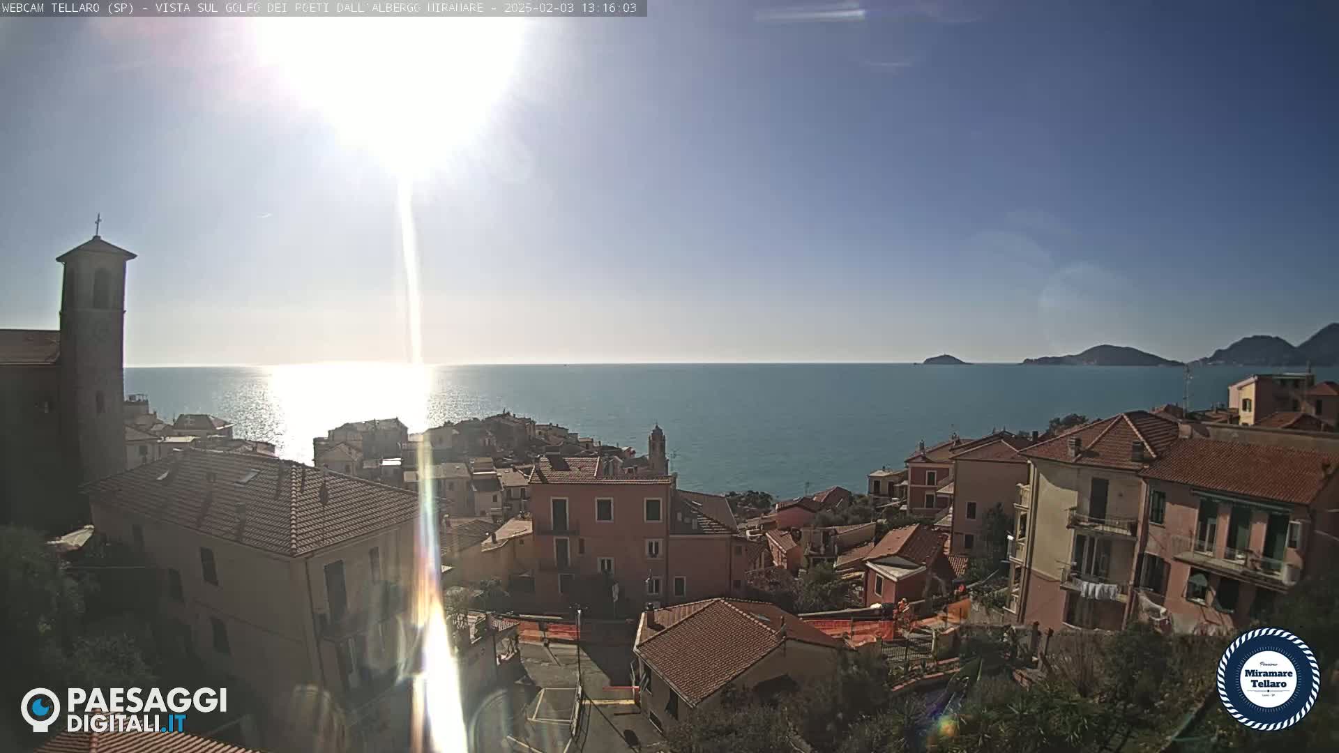 A sunlit coastal town with terracotta-roofed buildings overlooks a calm sea under a clear blue sky.