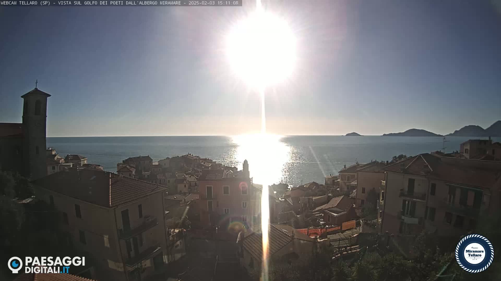 A coastal town with red-tiled roofs sits on a hillside overlooking a sunlit sea, under a bright, clear sky.