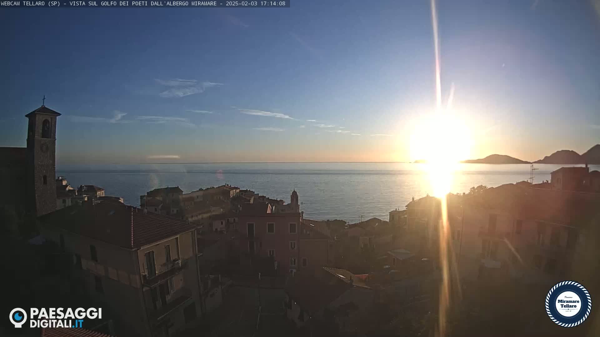 A coastal town with a church steeple is silhouetted against a setting sun over a calm sea.