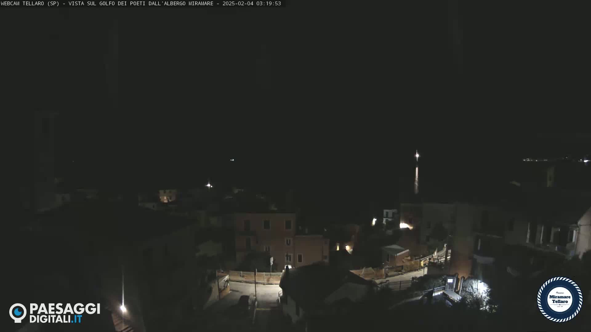 A nighttime, long shot of a coastal town overlooking dark water, with a few lights visible on buildings and streets.