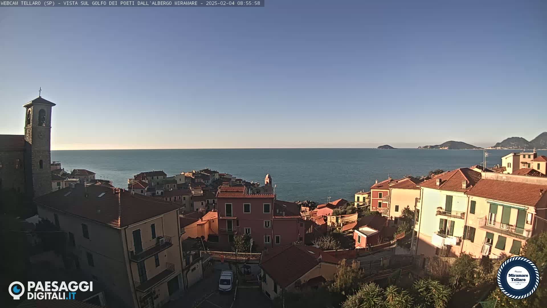 A coastal town with red-tiled roofs and a bell tower overlooks a calm sea under a clear blue sky.