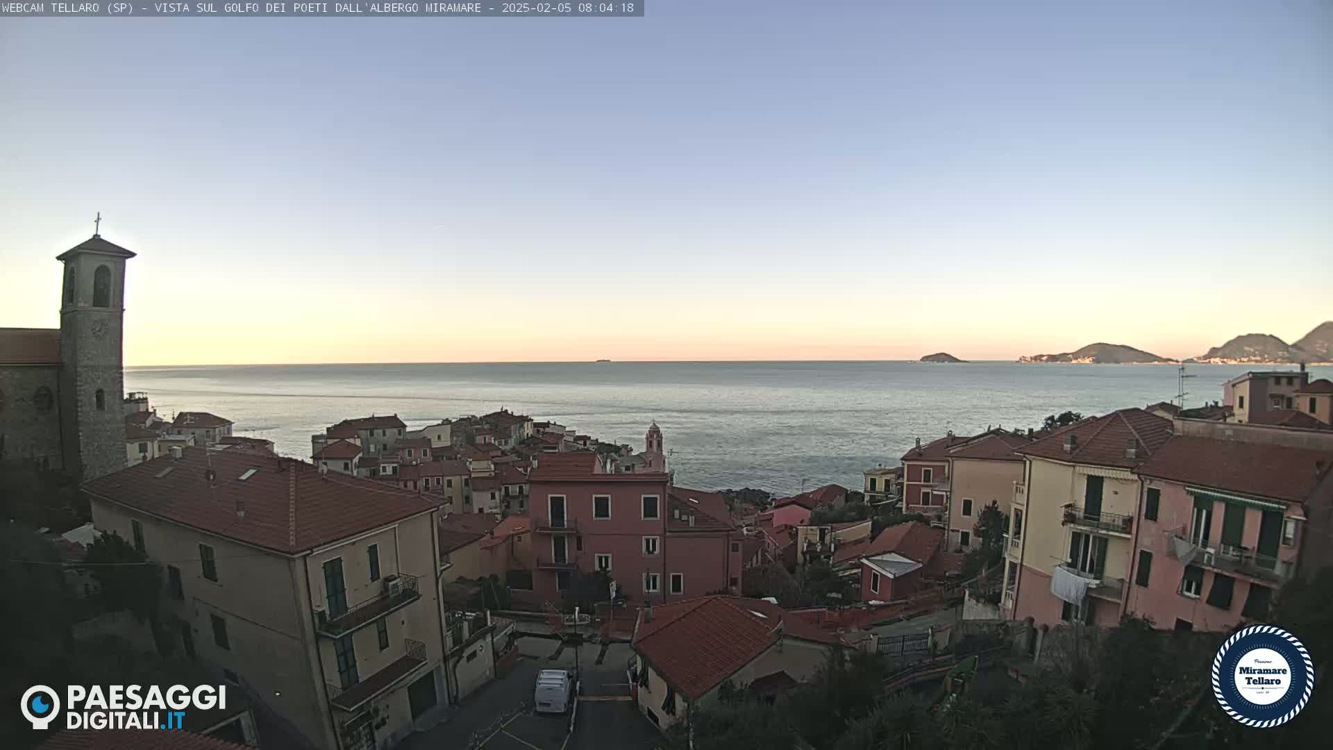 A coastal town with red-roofed buildings and a church steeple overlooks a calm sea under a clear, twilight sky.