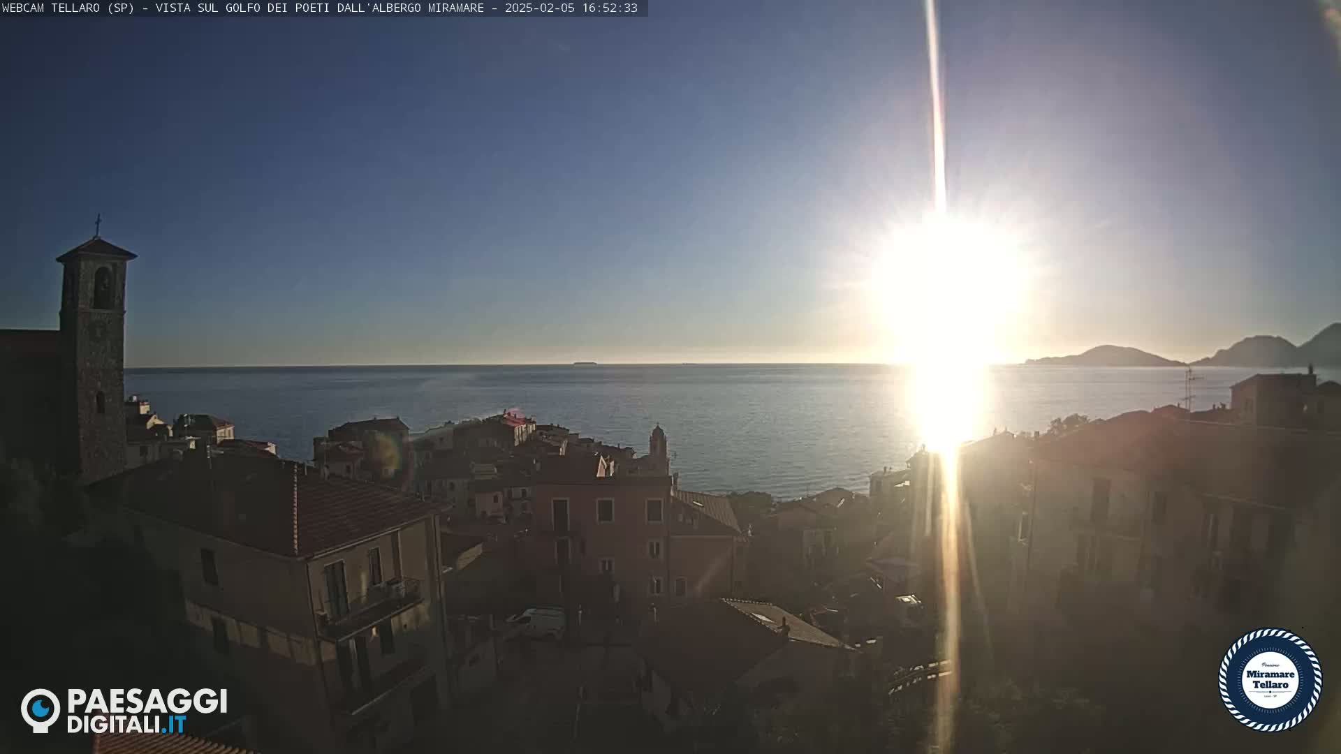 A coastal town with a bell tower is bathed in the bright light of the setting sun over a calm sea.