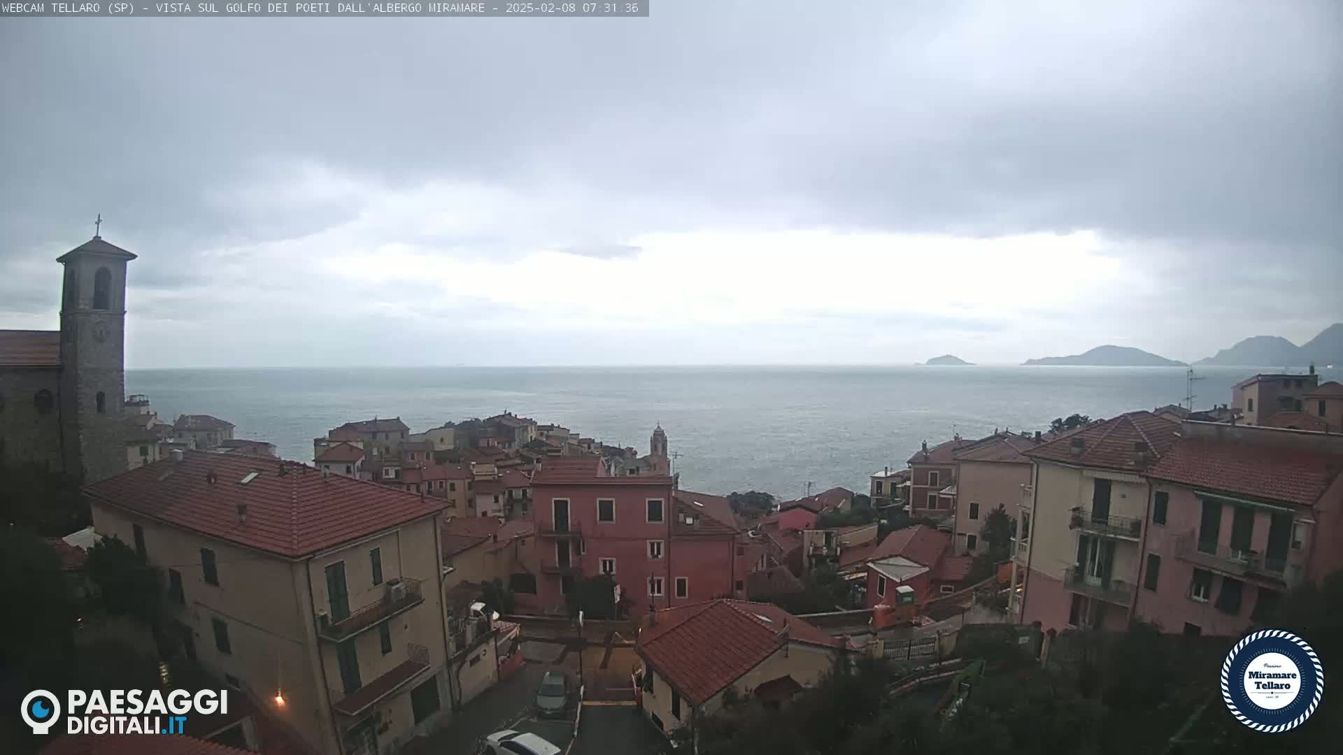 A coastal town with red-tiled roofs overlooks a calm sea under an overcast sky.