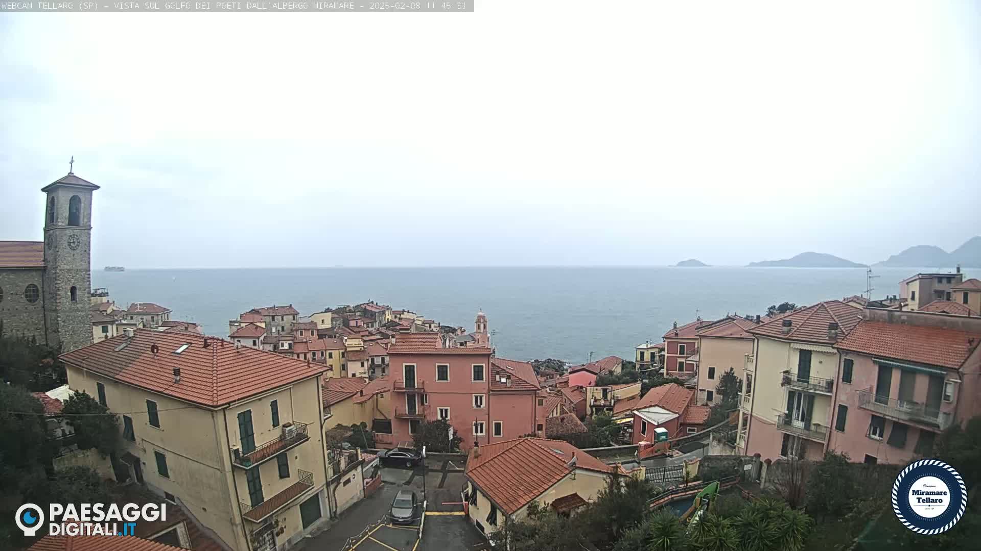 A coastal town with red-tiled roofs and a church overlooks a calm sea under an overcast sky.