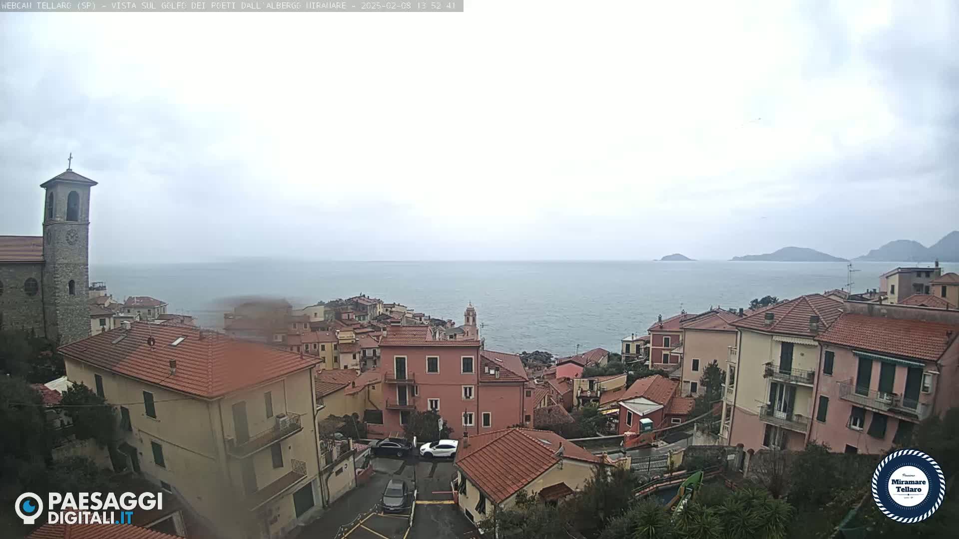 A coastal town with red-tiled roofs and a church steeple overlooks a calm sea under an overcast sky.