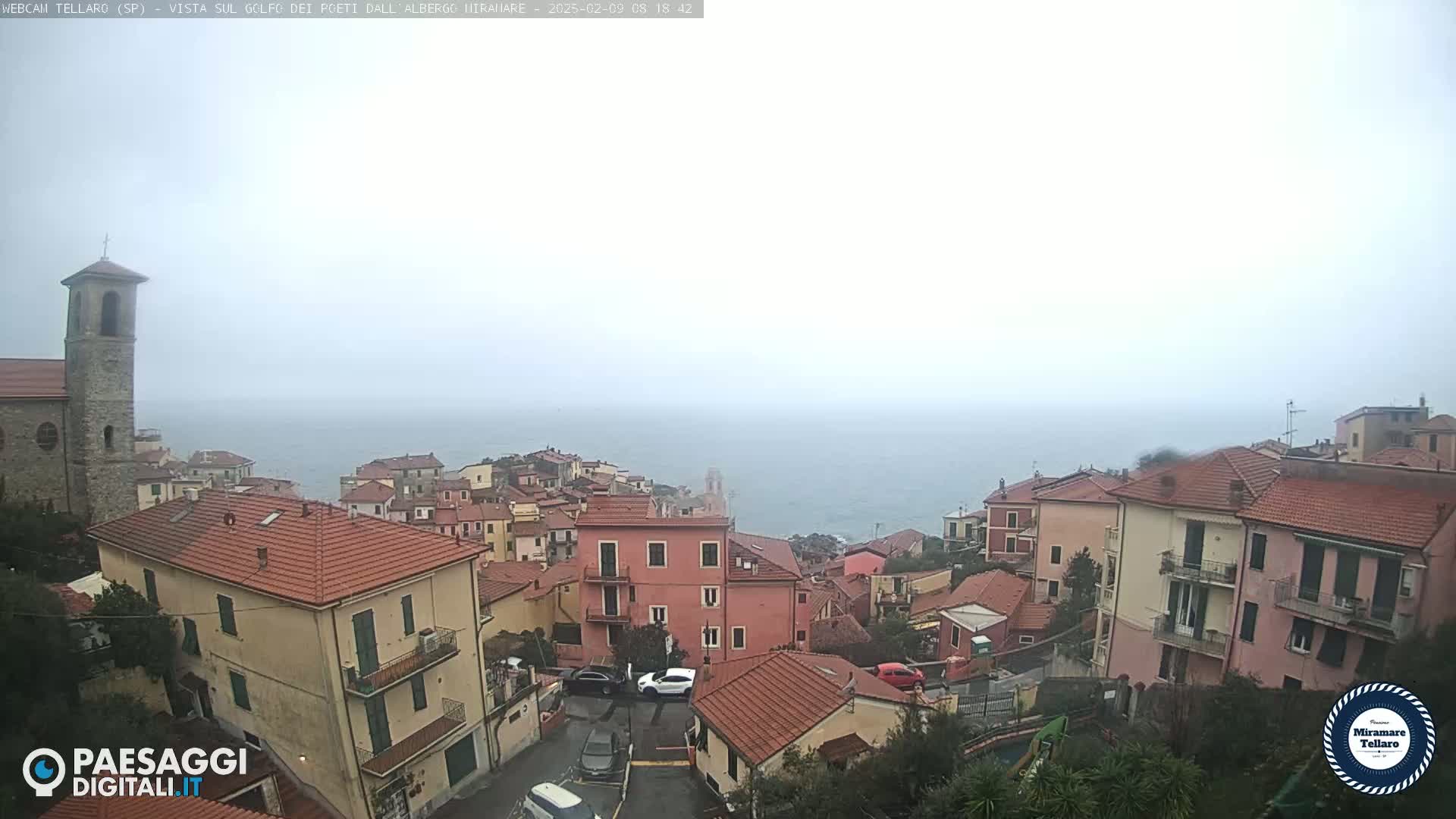 A foggy coastal town with red-roofed buildings overlooks a hazy sea, a church steeple visible in the background.