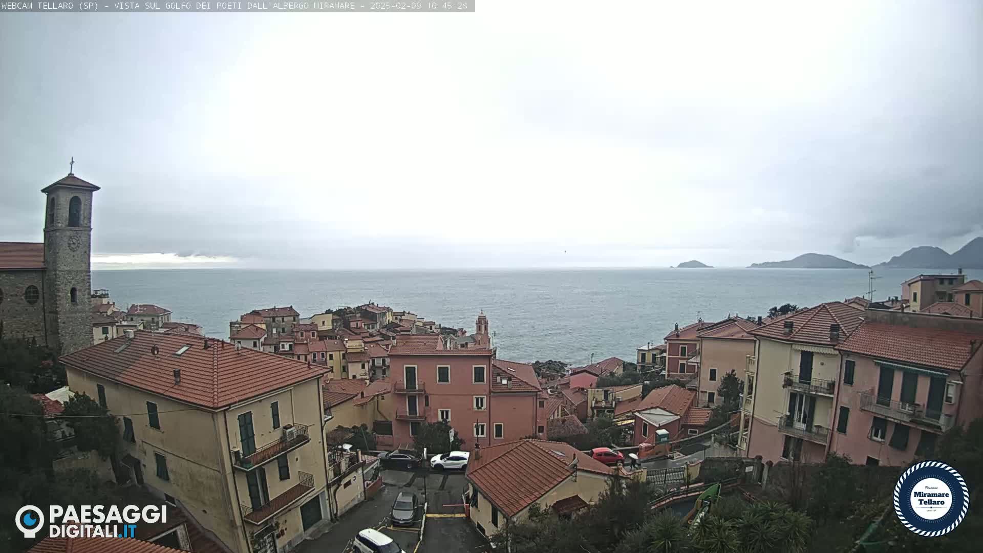 A coastal town with red-tiled roofs and a church overlooks a calm sea under an overcast sky.