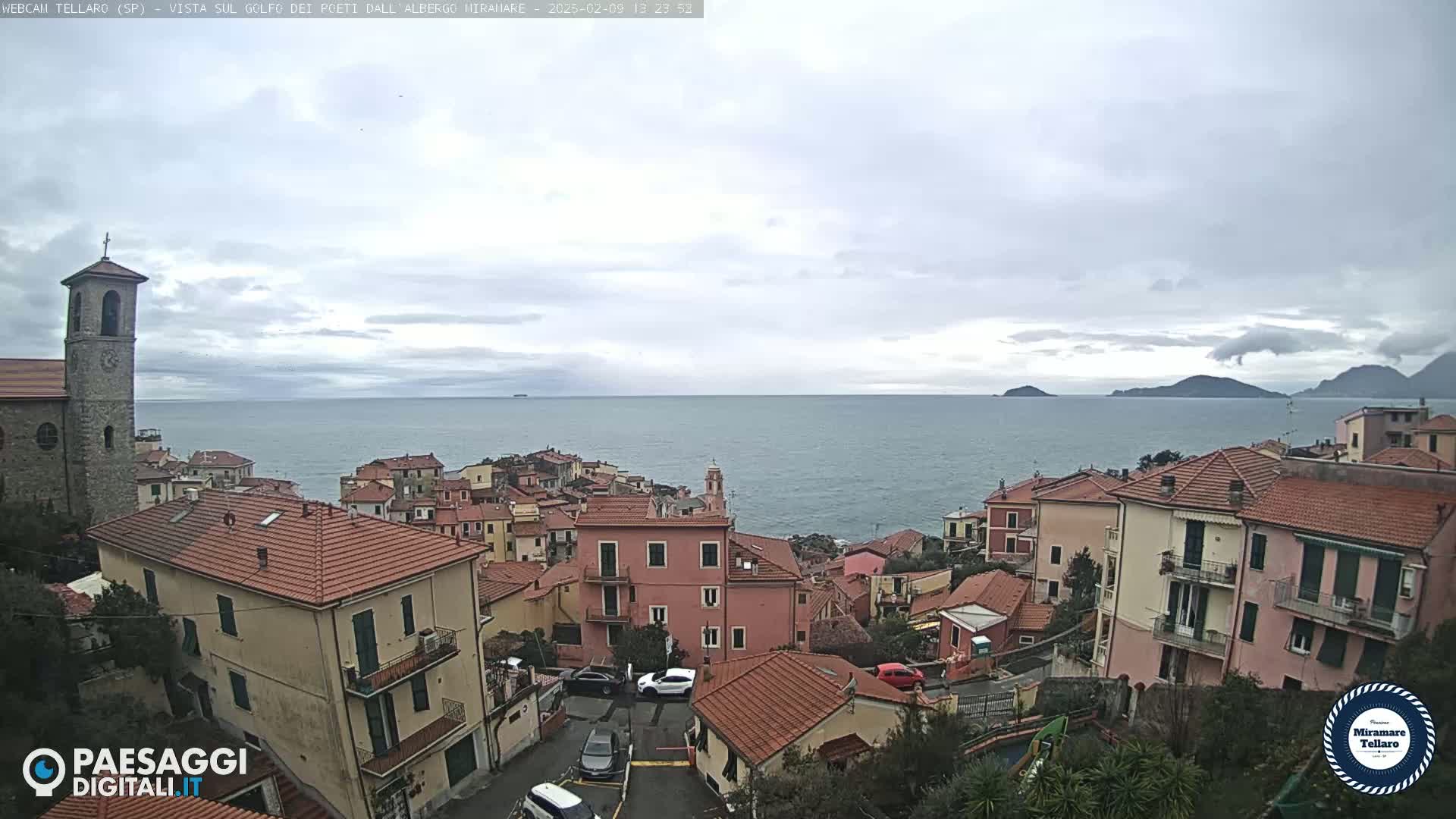A coastal town with red-tiled roofs overlooks a calm sea under a cloudy sky.