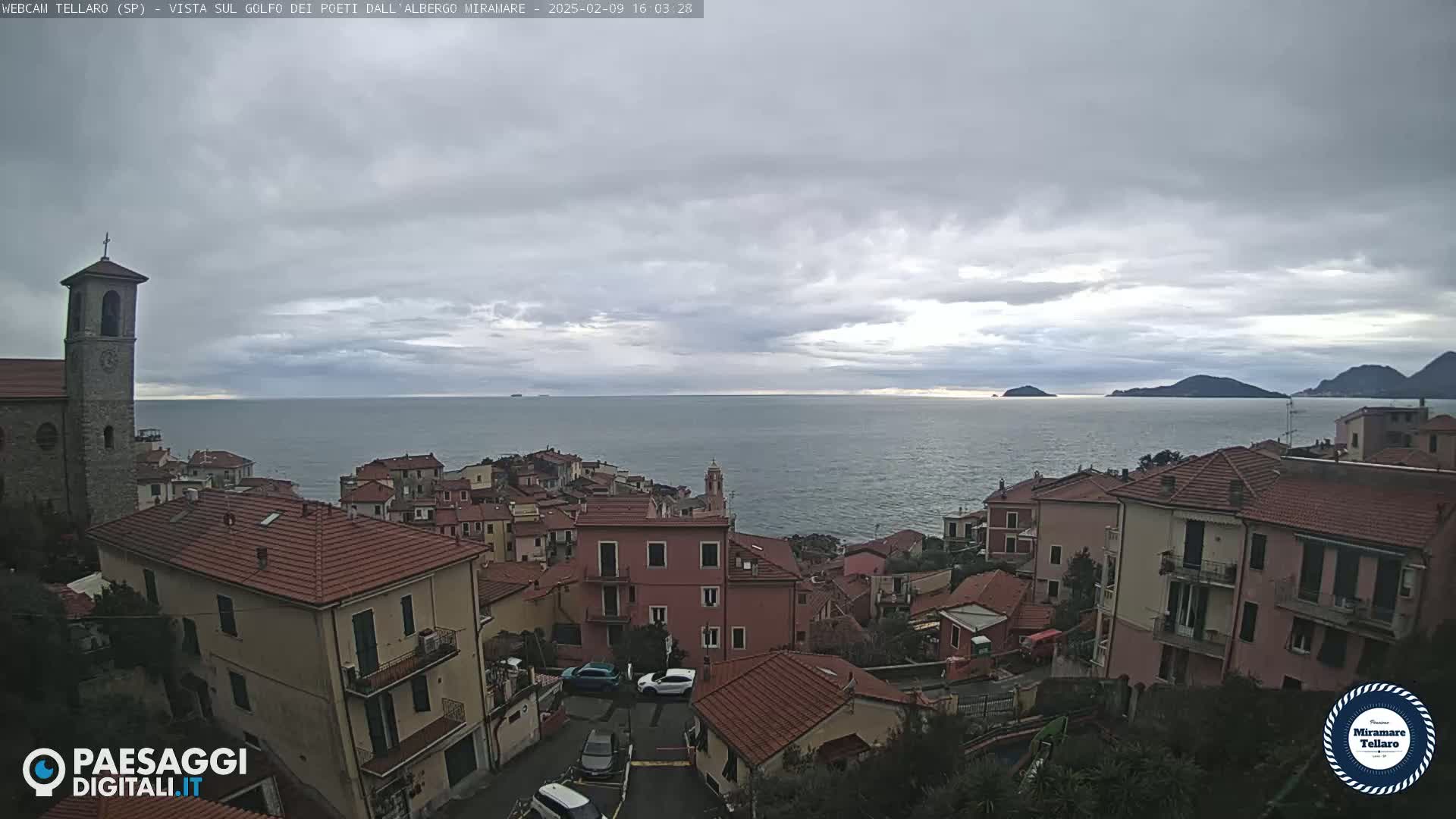 A coastal town with red-tiled roofs overlooks a calm sea under a cloudy sky.