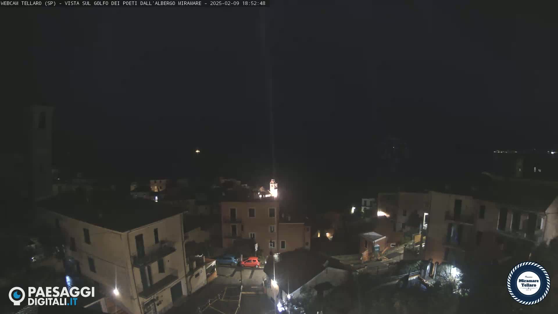 A nighttime, high-angle view of a small town, possibly coastal, with dimly lit buildings and a few cars parked in a small square.