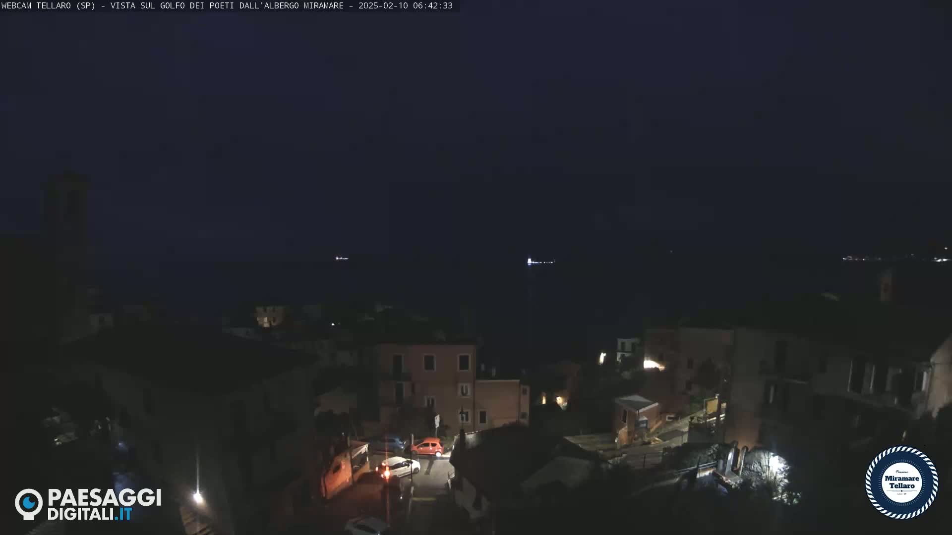 A nighttime, aerial view of a coastal town with a few cars parked on a street, overlooking a dark body of water.