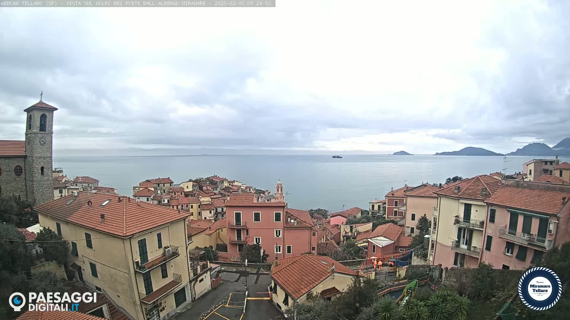 A coastal town with red-tiled roofs overlooks a calm sea under a cloudy sky.