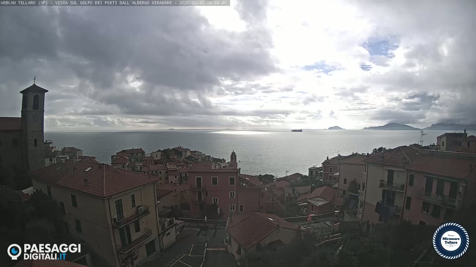A coastal town with red-tiled roofs overlooks a calm sea under a mostly cloudy sky.