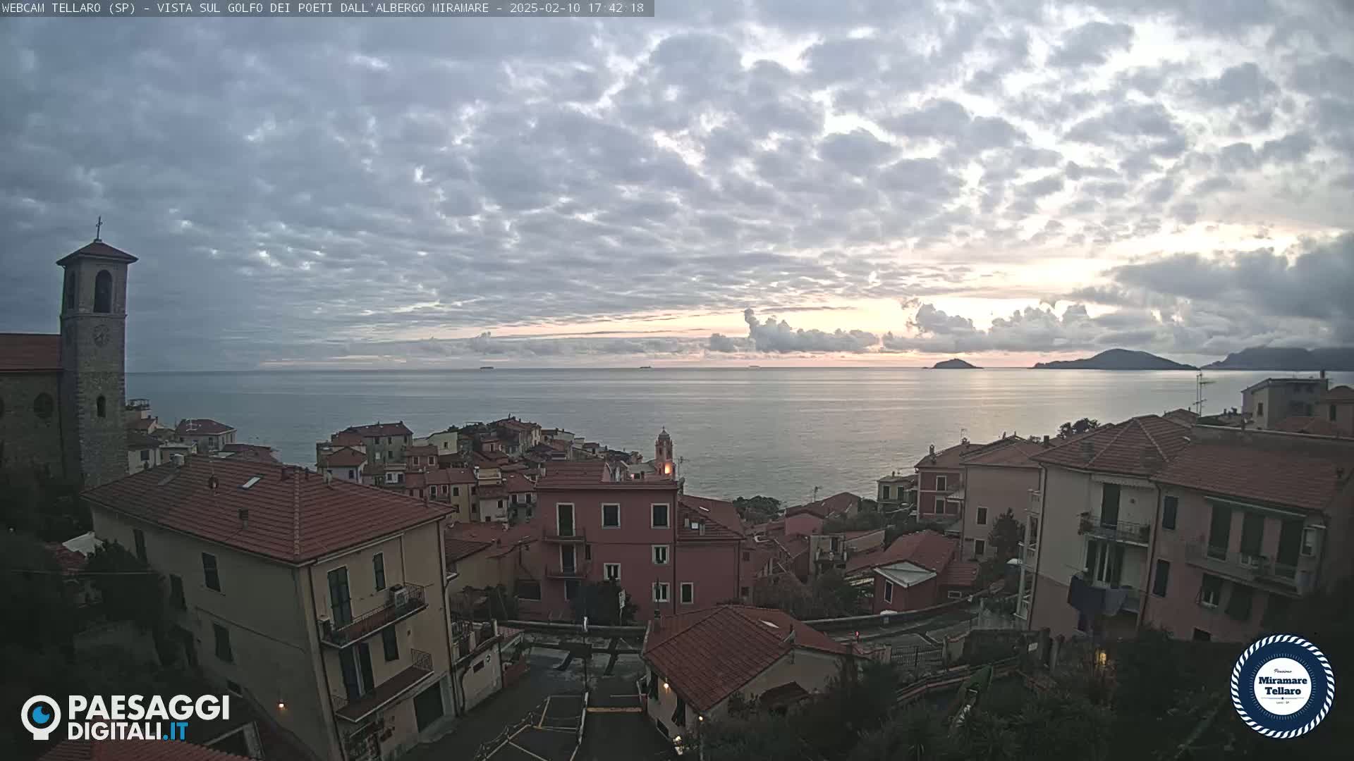 A coastal town with red-tiled roofs overlooks a calm sea under a mostly cloudy, dusky sky.