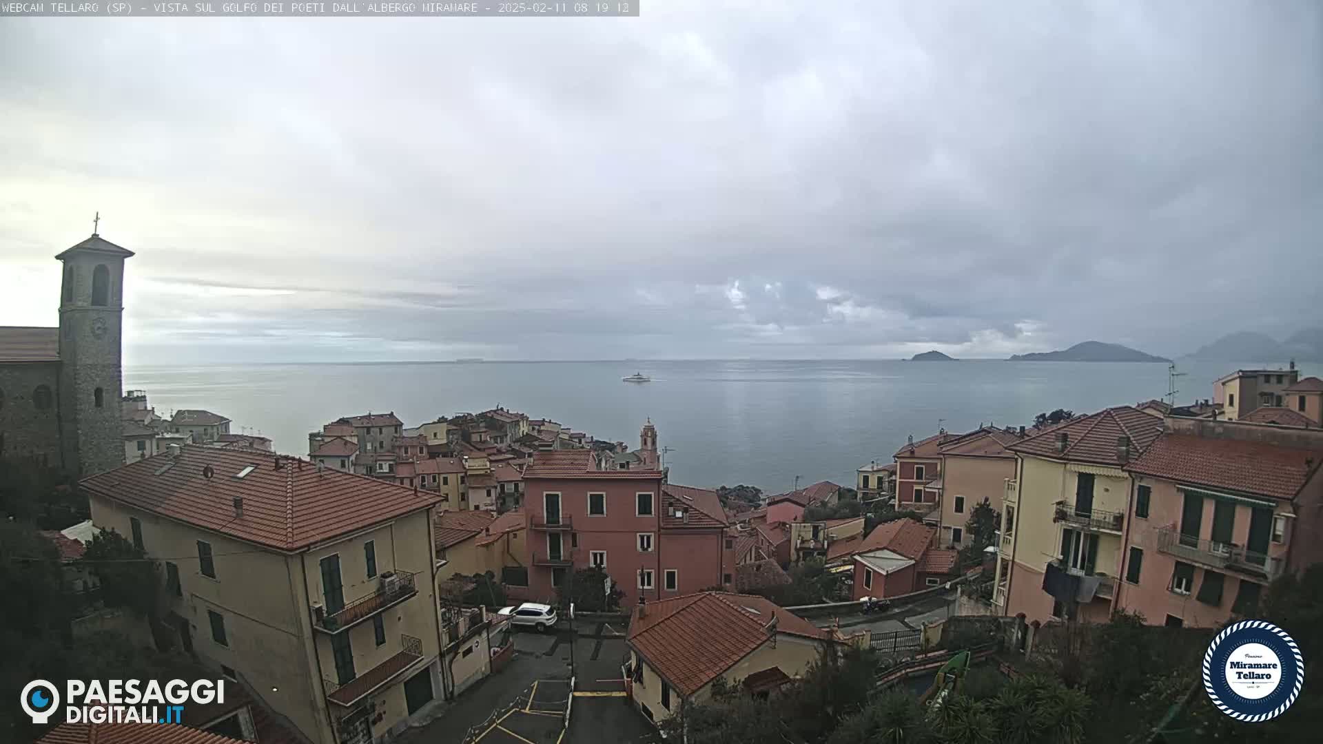 A coastal town with red-tiled roofs overlooks a calm sea under a cloudy sky.