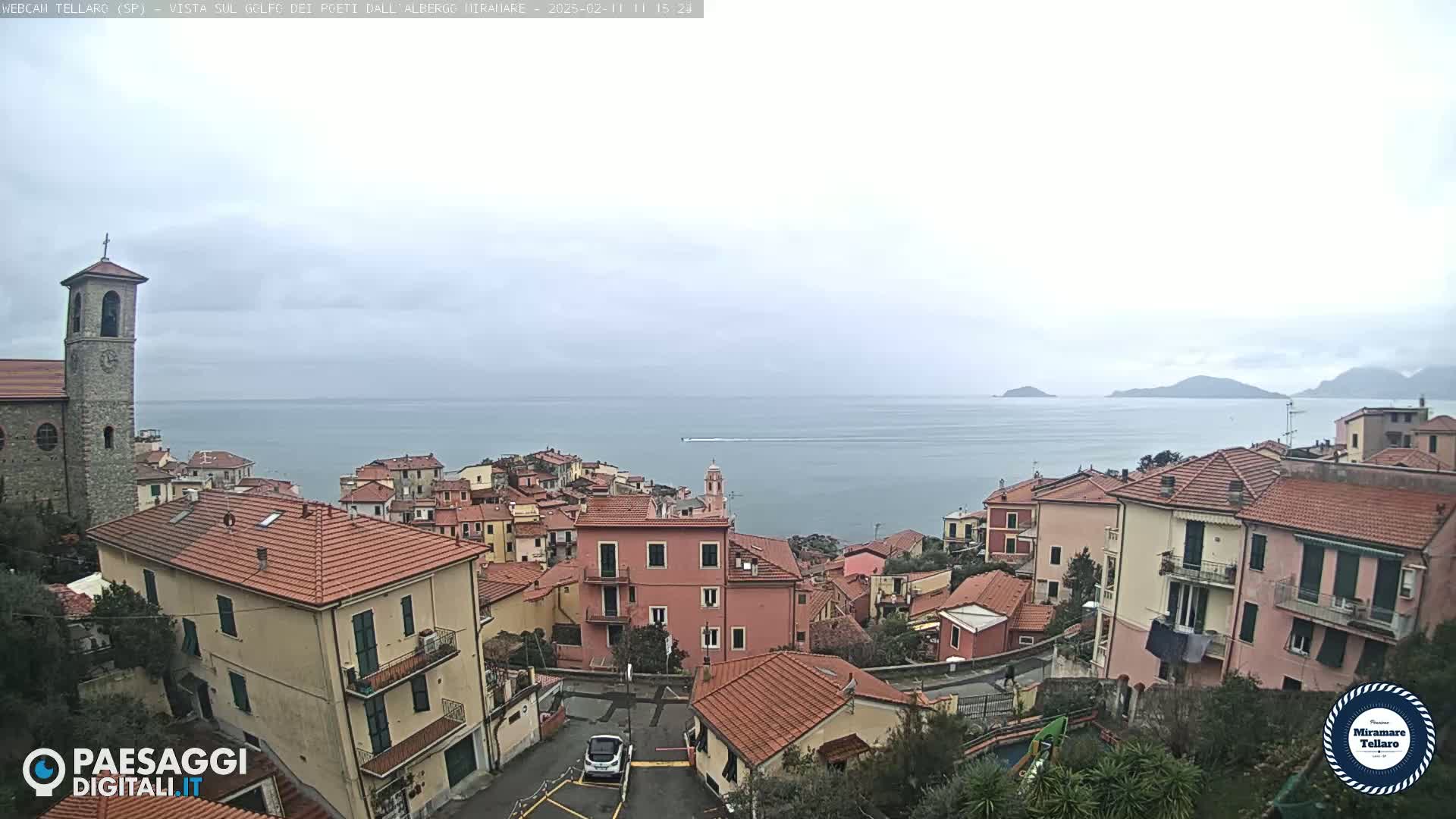 An overcast day reveals a coastal village with red-tiled roofs, a church bell tower, and a calm sea extending to distant hills.