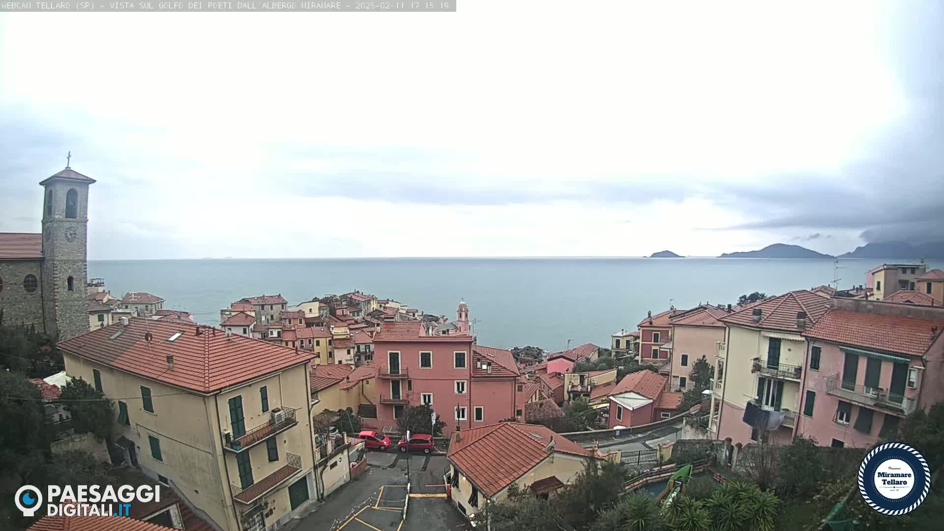 A coastal town with red-tiled roofs and a church overlooks a calm sea under a cloudy sky.