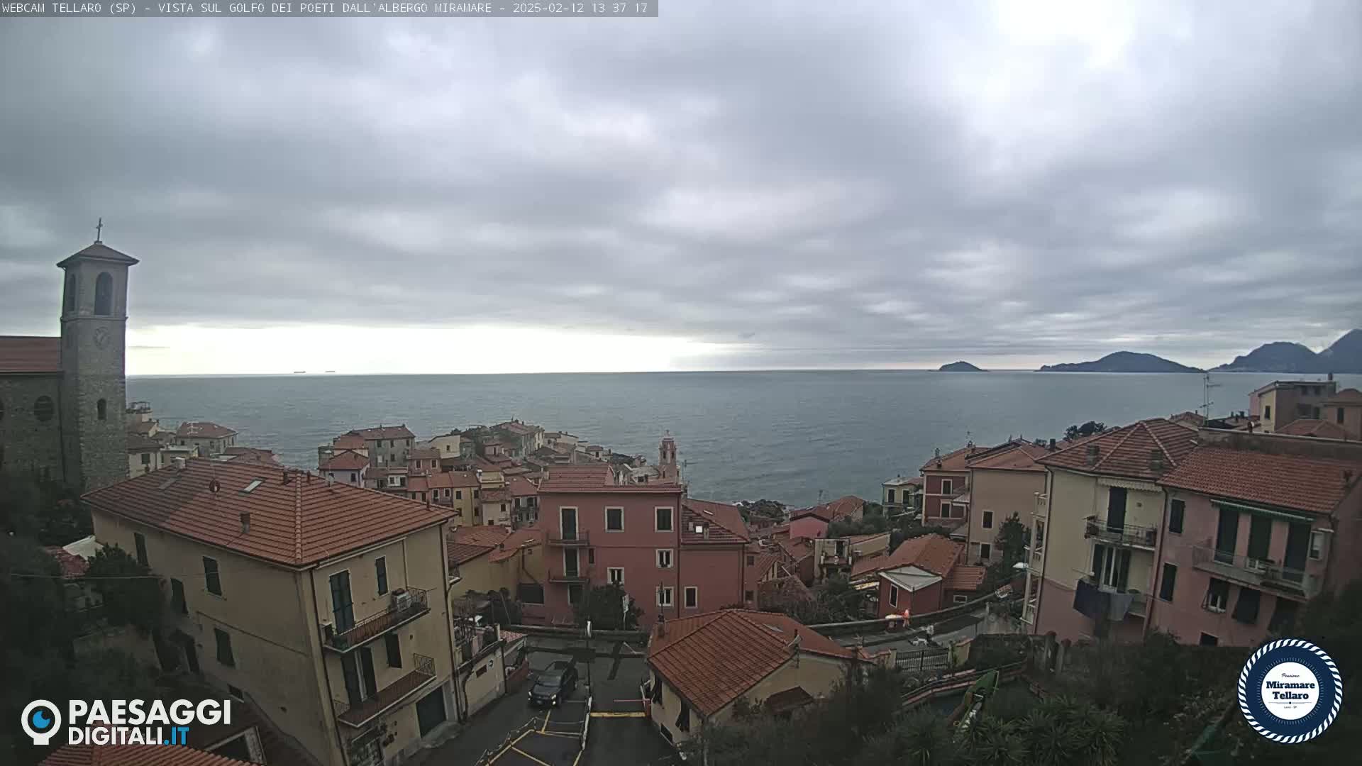 A coastal town with red-tiled roofs overlooks a calm sea under a cloudy sky.